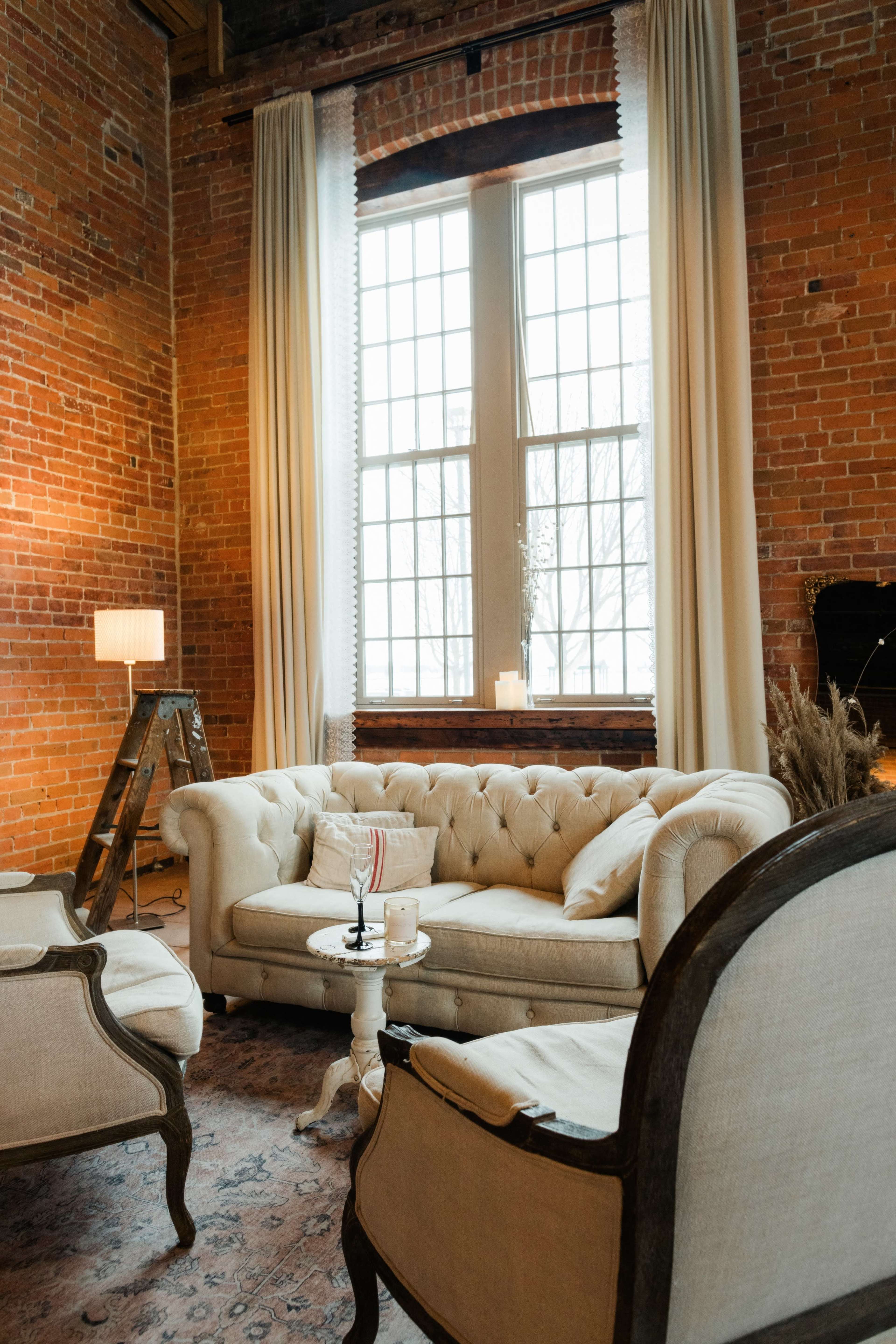 A cozy living area featuring a tufted beige sofa, two upholstered chairs, a small table, and large windows with white curtains against a backdrop of exposed brick walls.