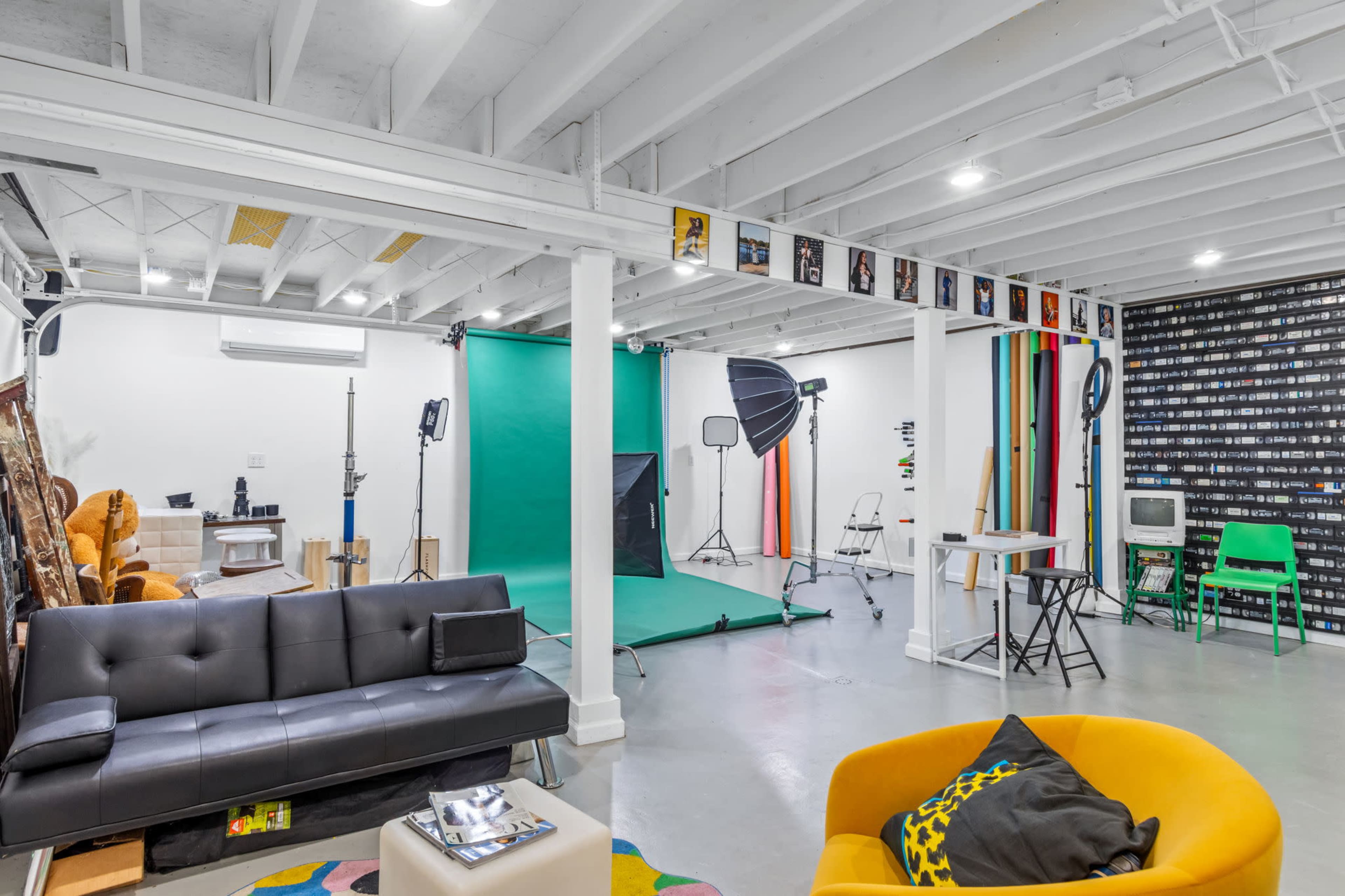 The image shows a photo studio with a green backdrop, studio lighting, and various props arranged around a seating area with modern furniture.