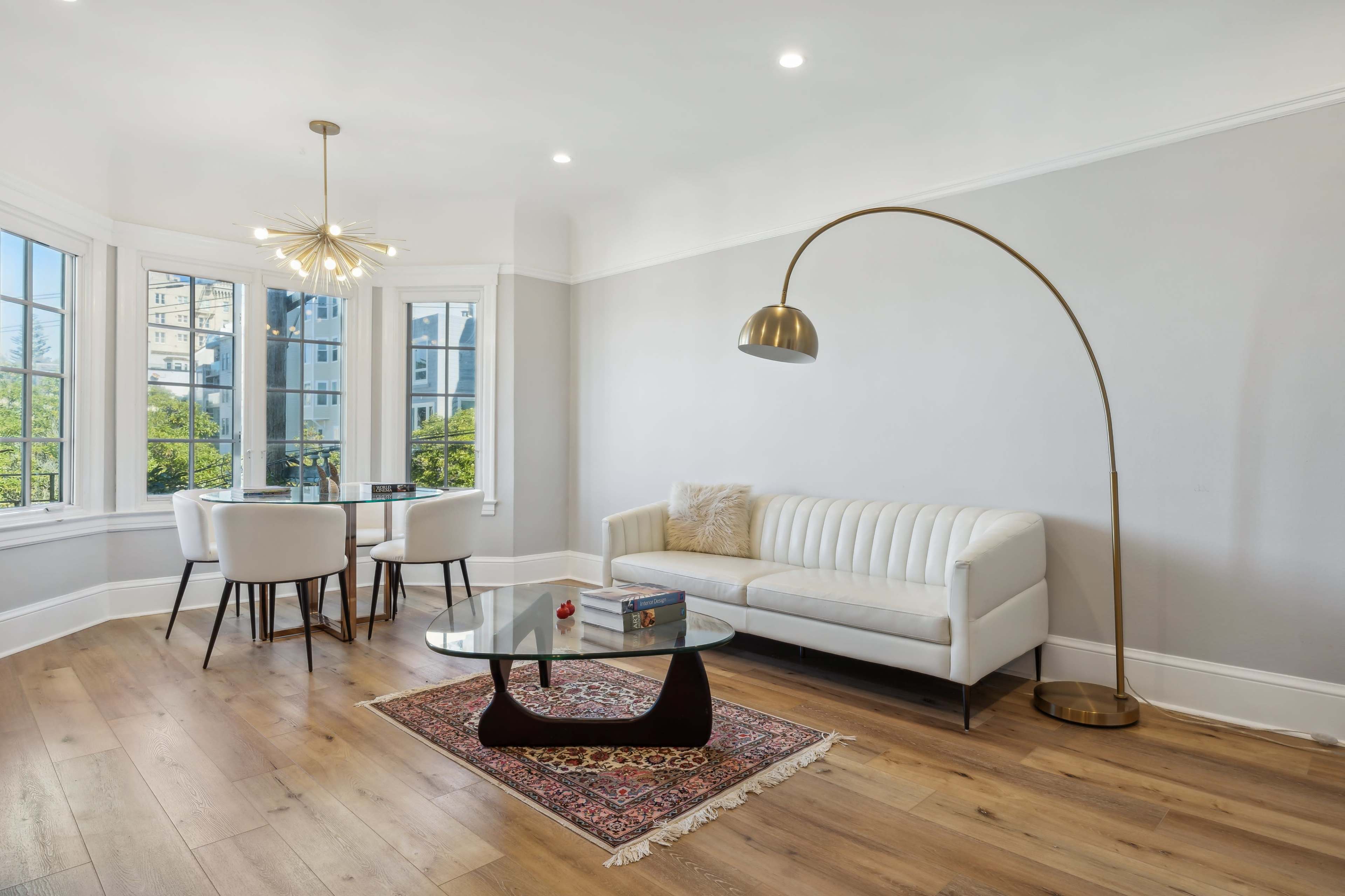 A modern living space features a white sofa, a glass coffee table on a decorative rug, a dining table with four chairs, and a tall floor lamp, all set against large windows.