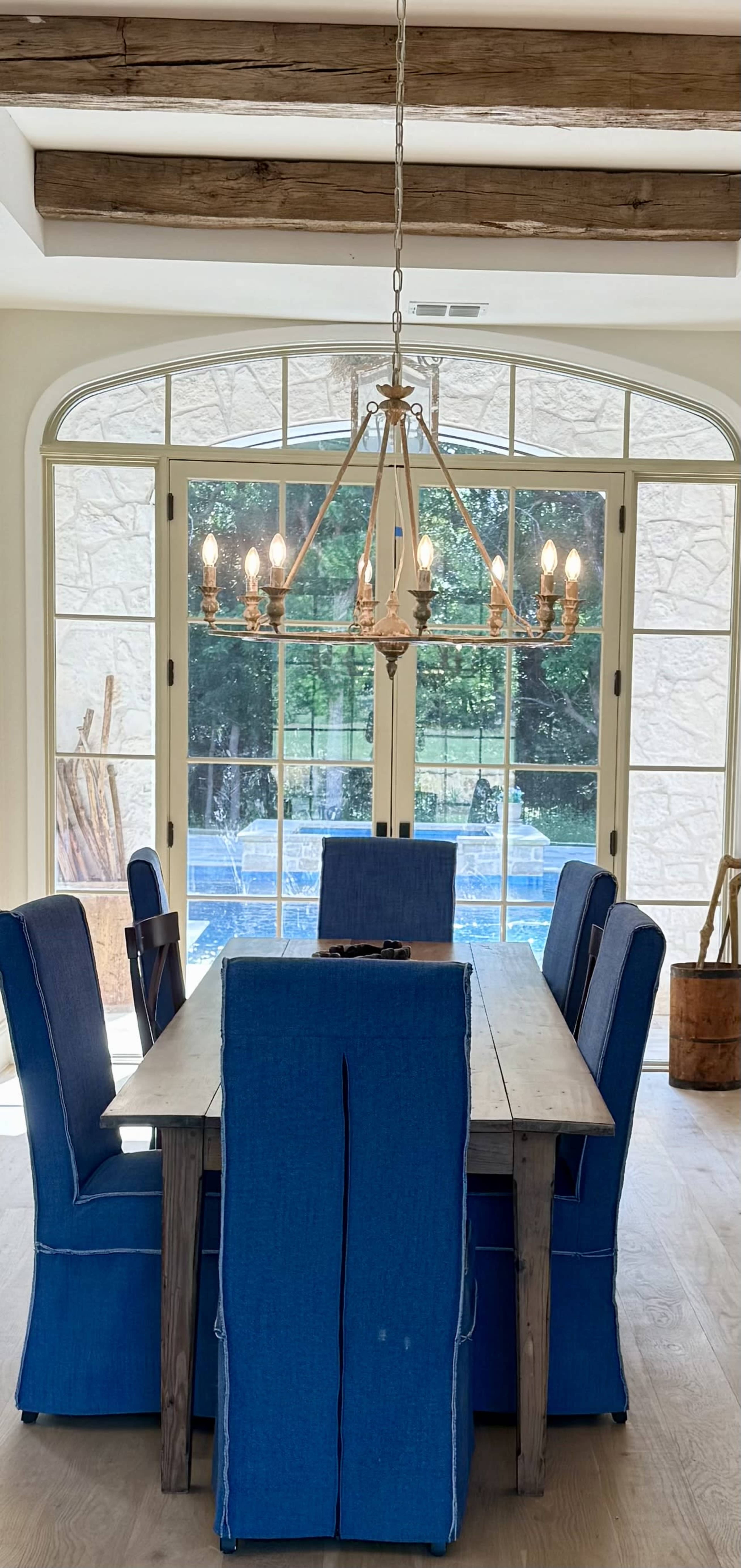 A dining room features a large wooden table surrounded by blue upholstered chairs and a chandelier hanging from a beamed ceiling, with large windows overlooking a pool area.