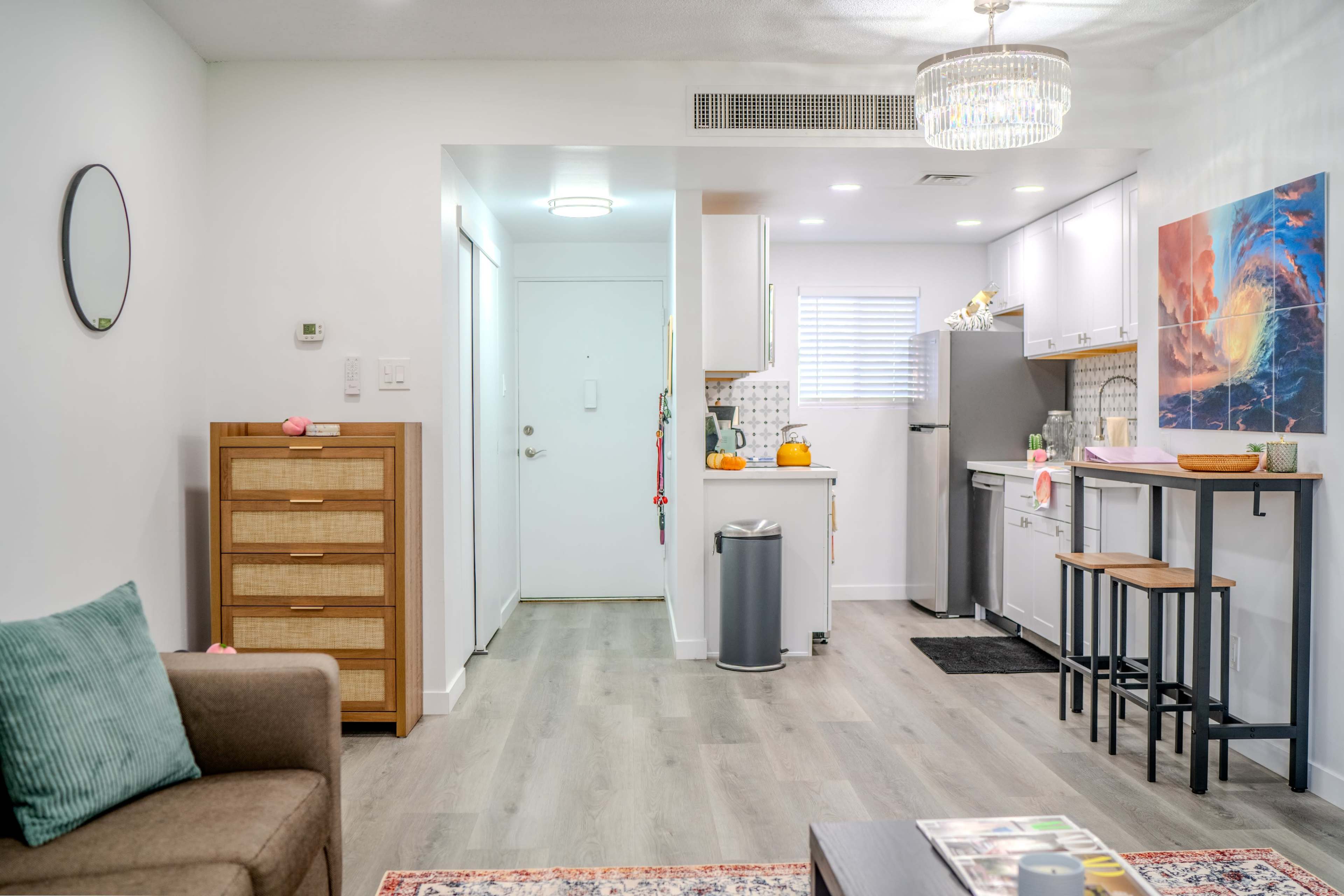 The image shows a modern, well-lit living space featuring a small kitchen, a dining area with bar stools, and a cozy seating area with a sofa and decorative rug.