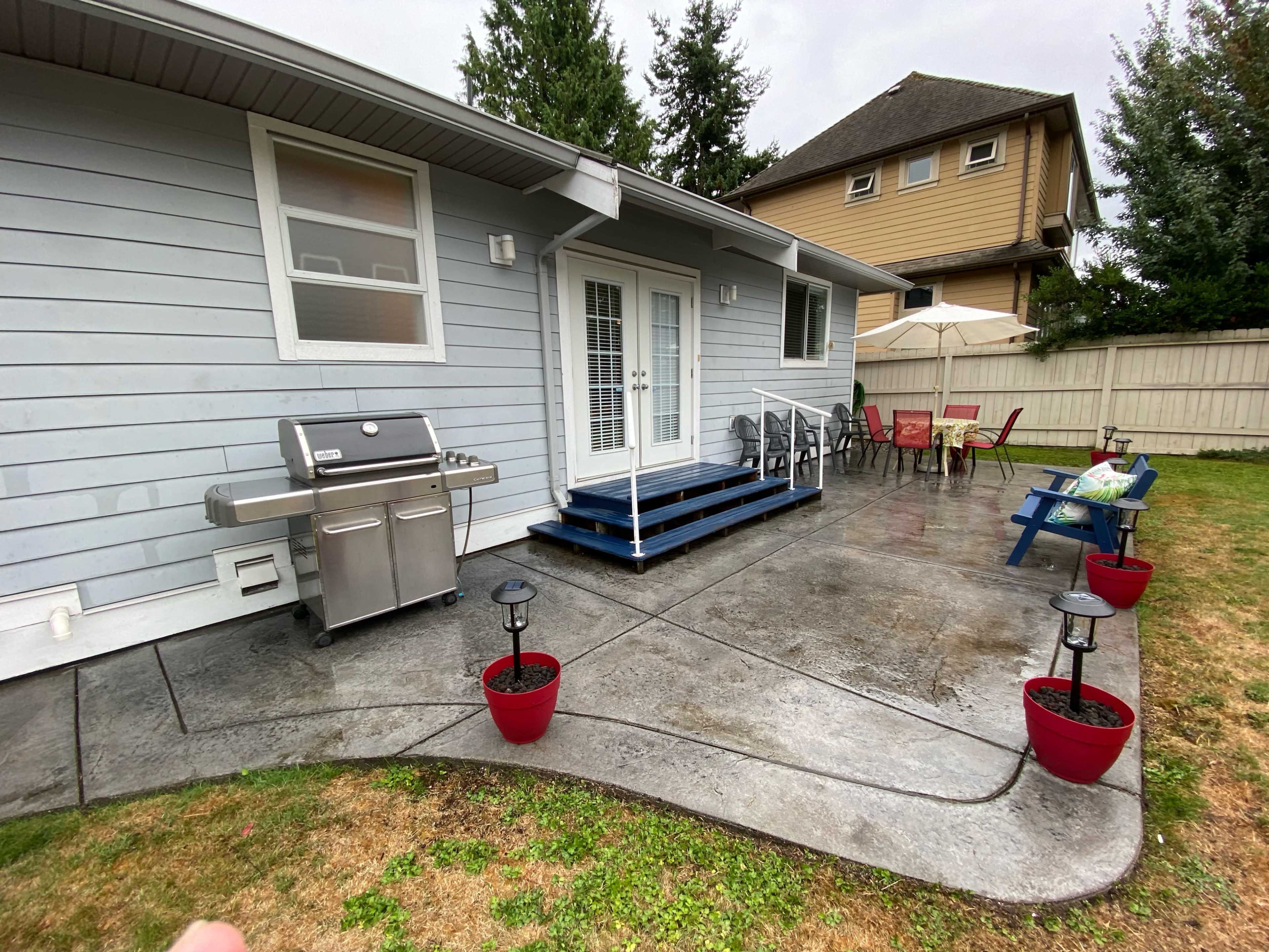 A gray house with a patio featuring a BBQ grill, outdoor seating, and potted plants on a rainy day.