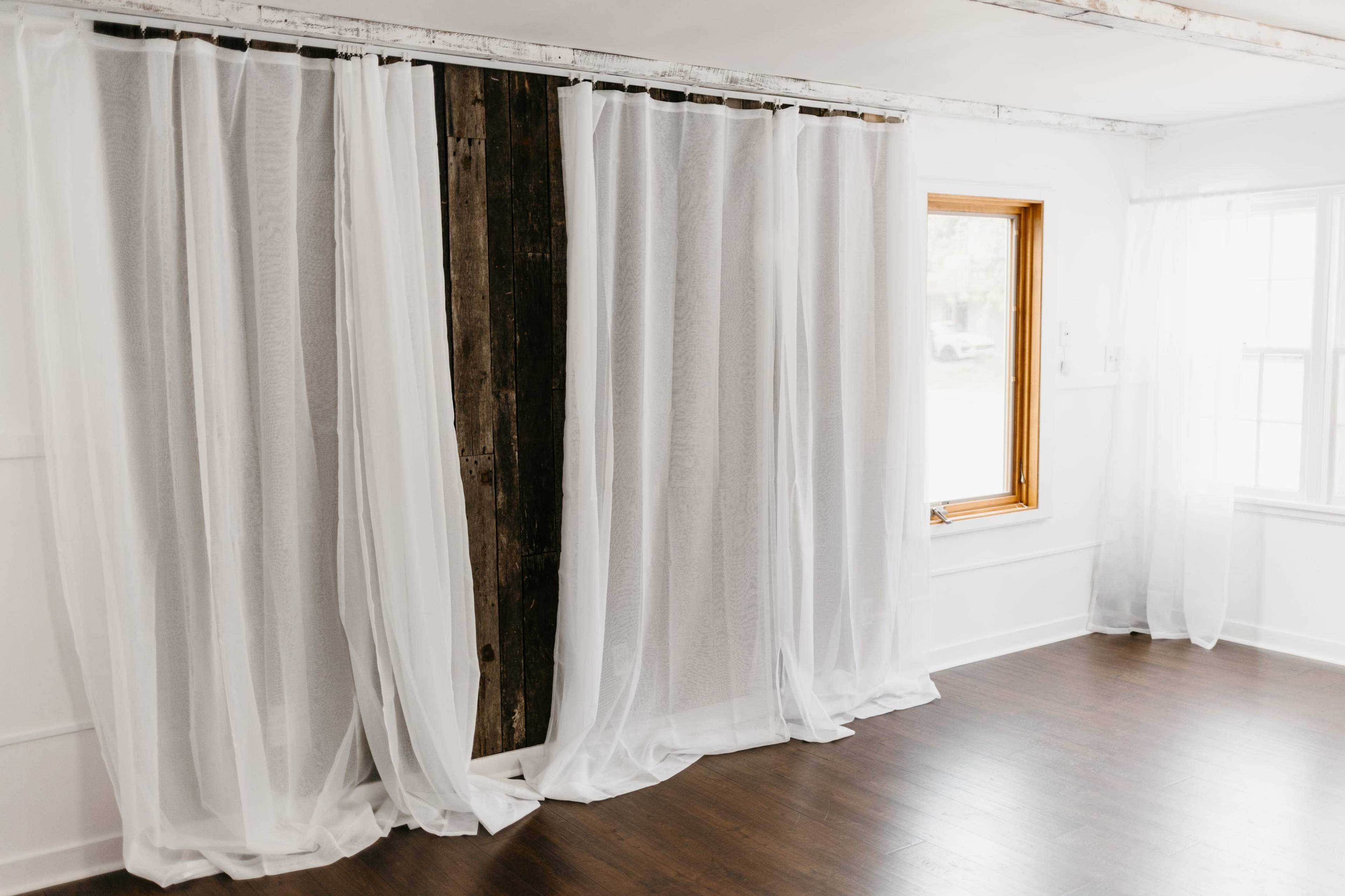Sheer white curtains drape elegantly over a wooden beam, framing a window in a bright, minimalist room with wooden flooring.