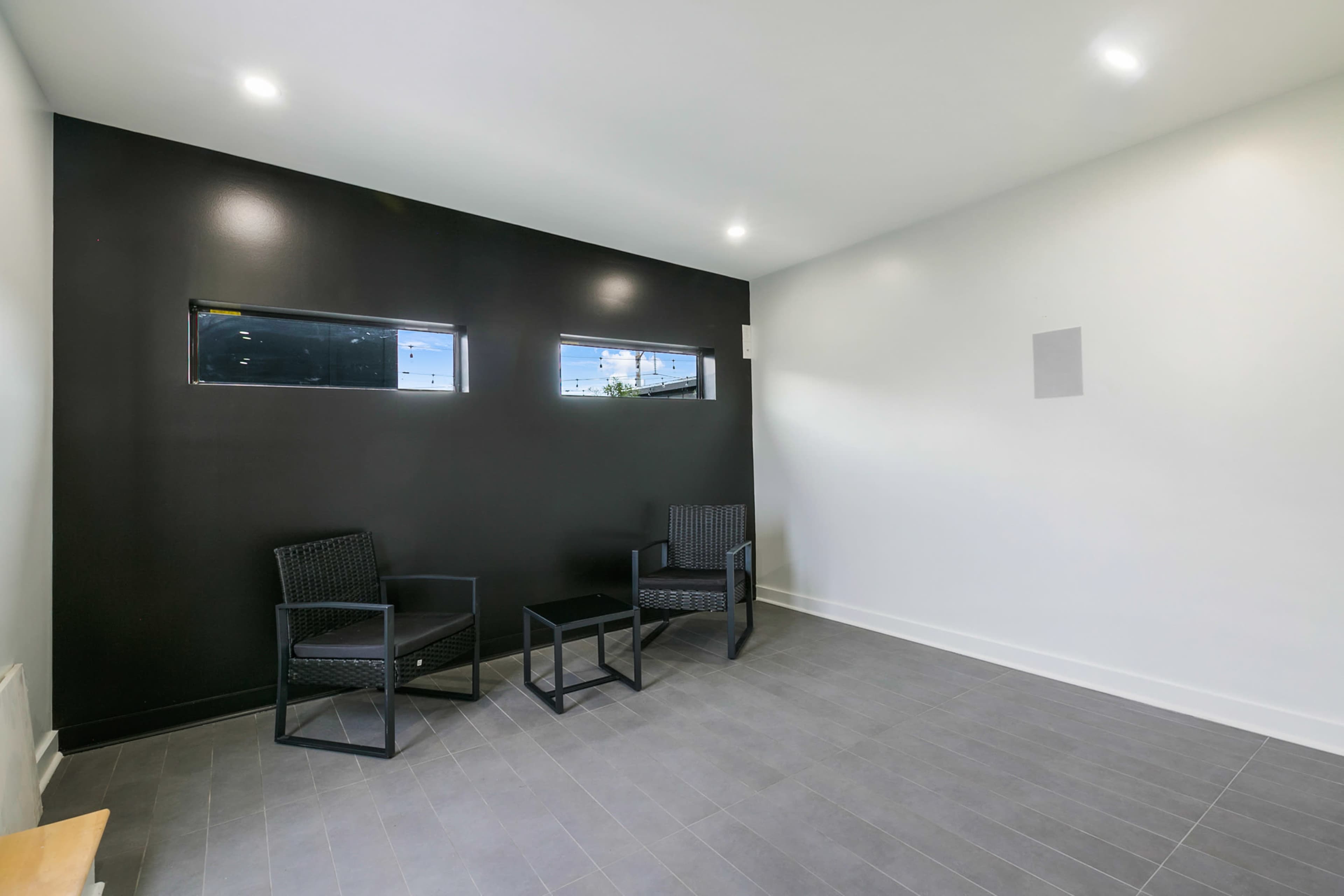 The image shows a minimalistic room with two black wicker chairs and a small table, featuring a dark wall with two narrow windows.