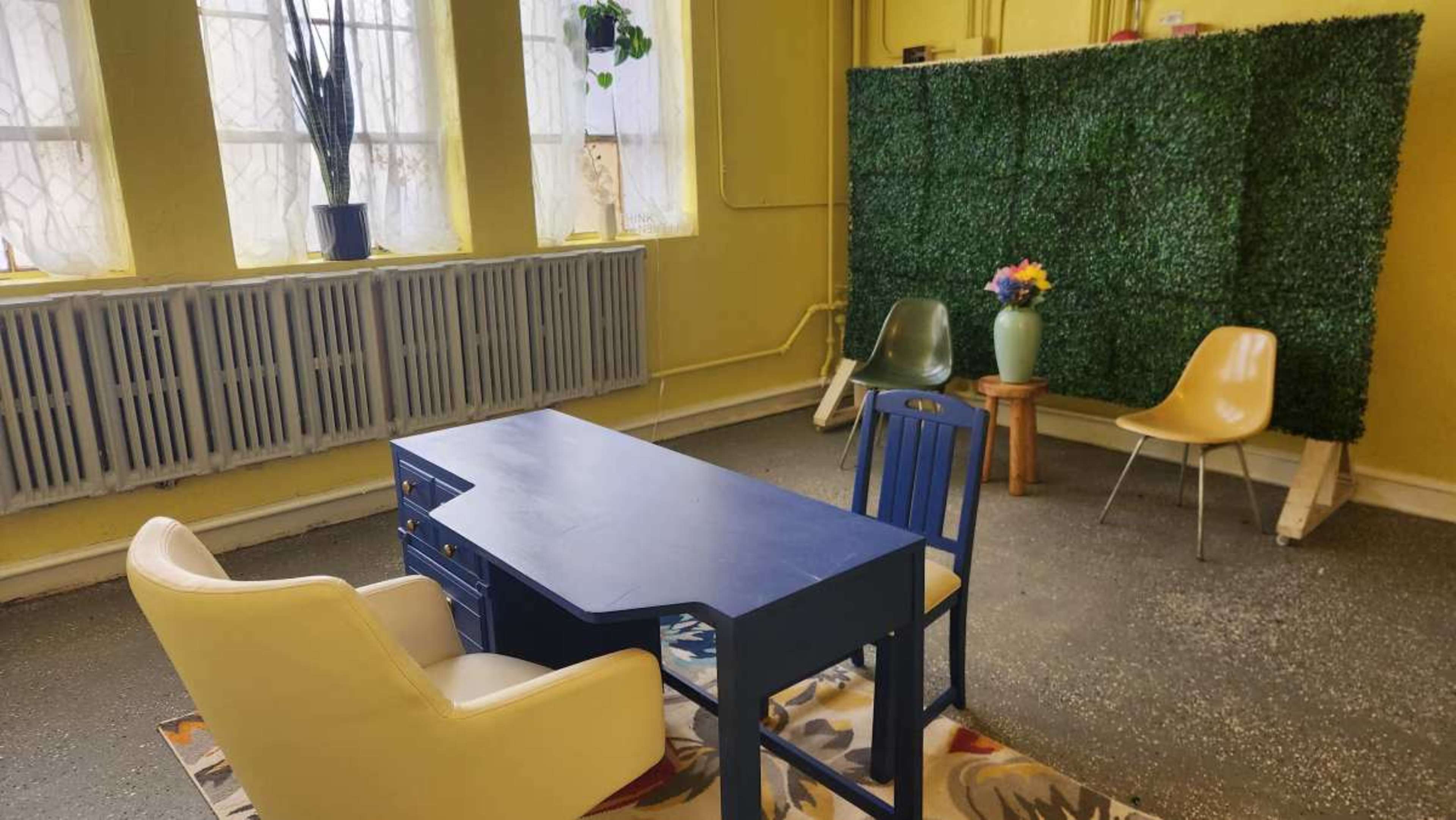The image shows a small room with yellow walls, featuring a blue desk and two chairs, alongside a green hedge wall and a vase of flowers on a pedestal.