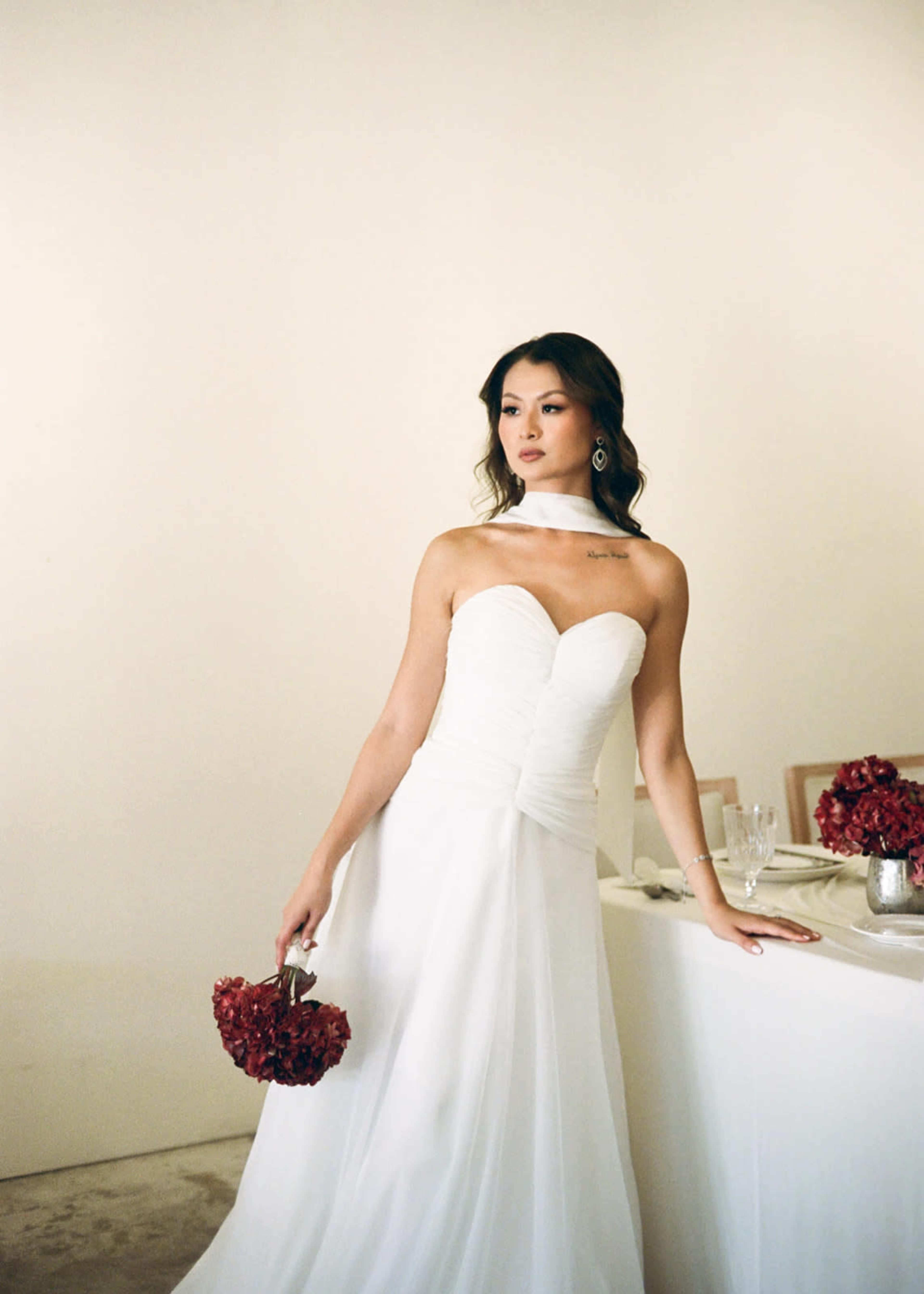 A woman in a white wedding dress poses elegantly with a bouquet of red flowers beside a table set for a celebration.