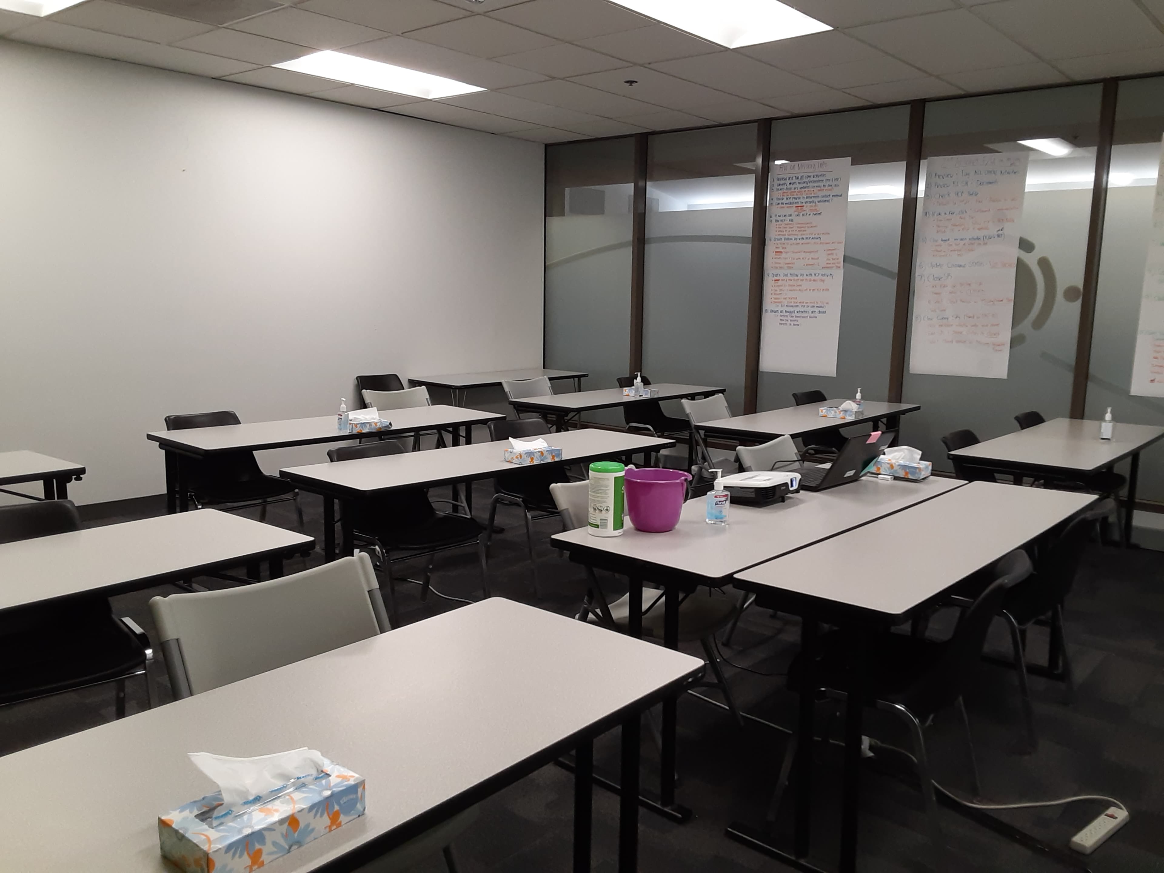A classroom setting with several empty tables and chairs, featuring supplies like tissues and water bottles on the surfaces.