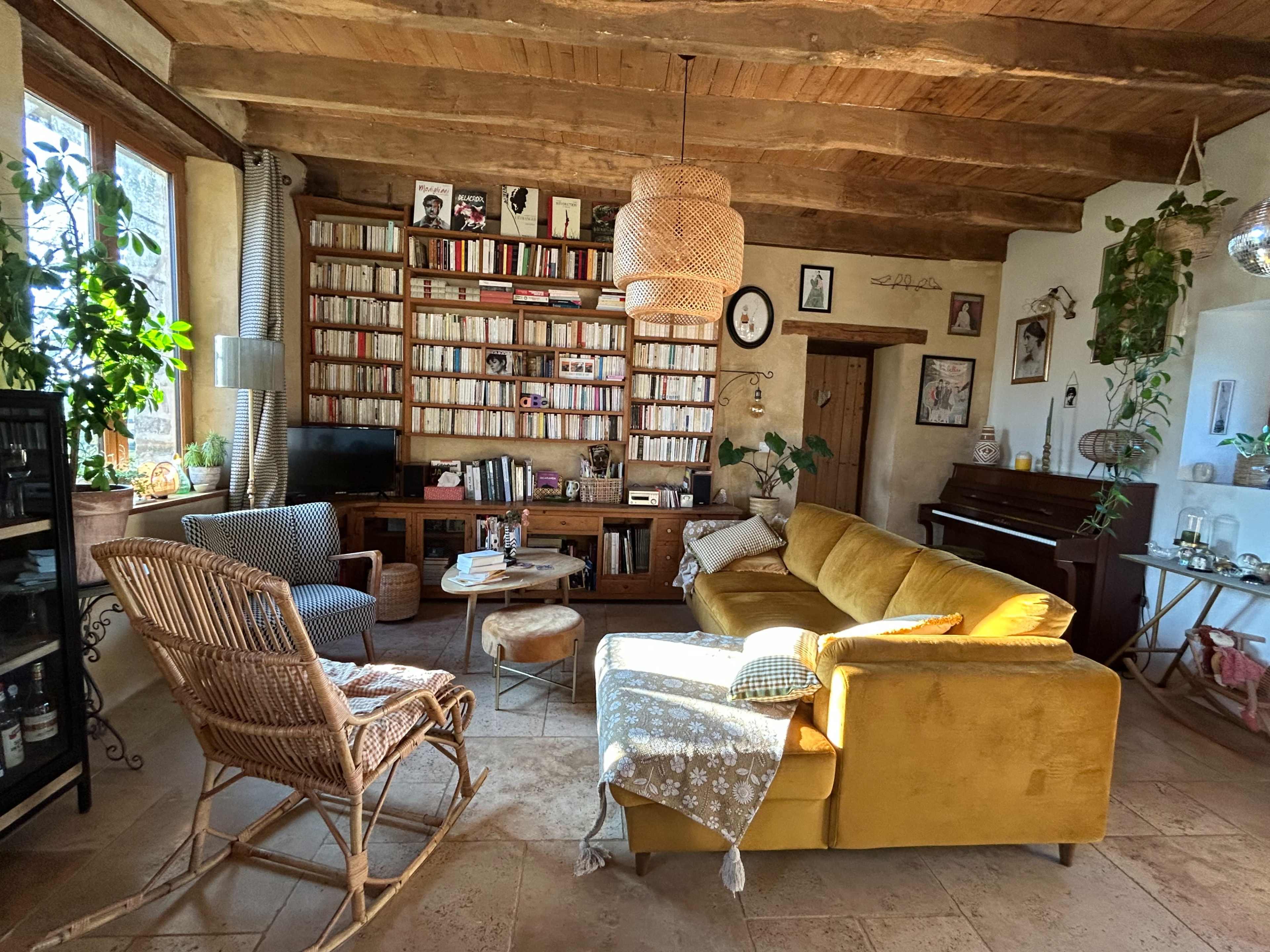 The image shows a cozy living room with wooden beams, a yellow sofa, a wicker chair, a bookshelf filled with books, and a piano.