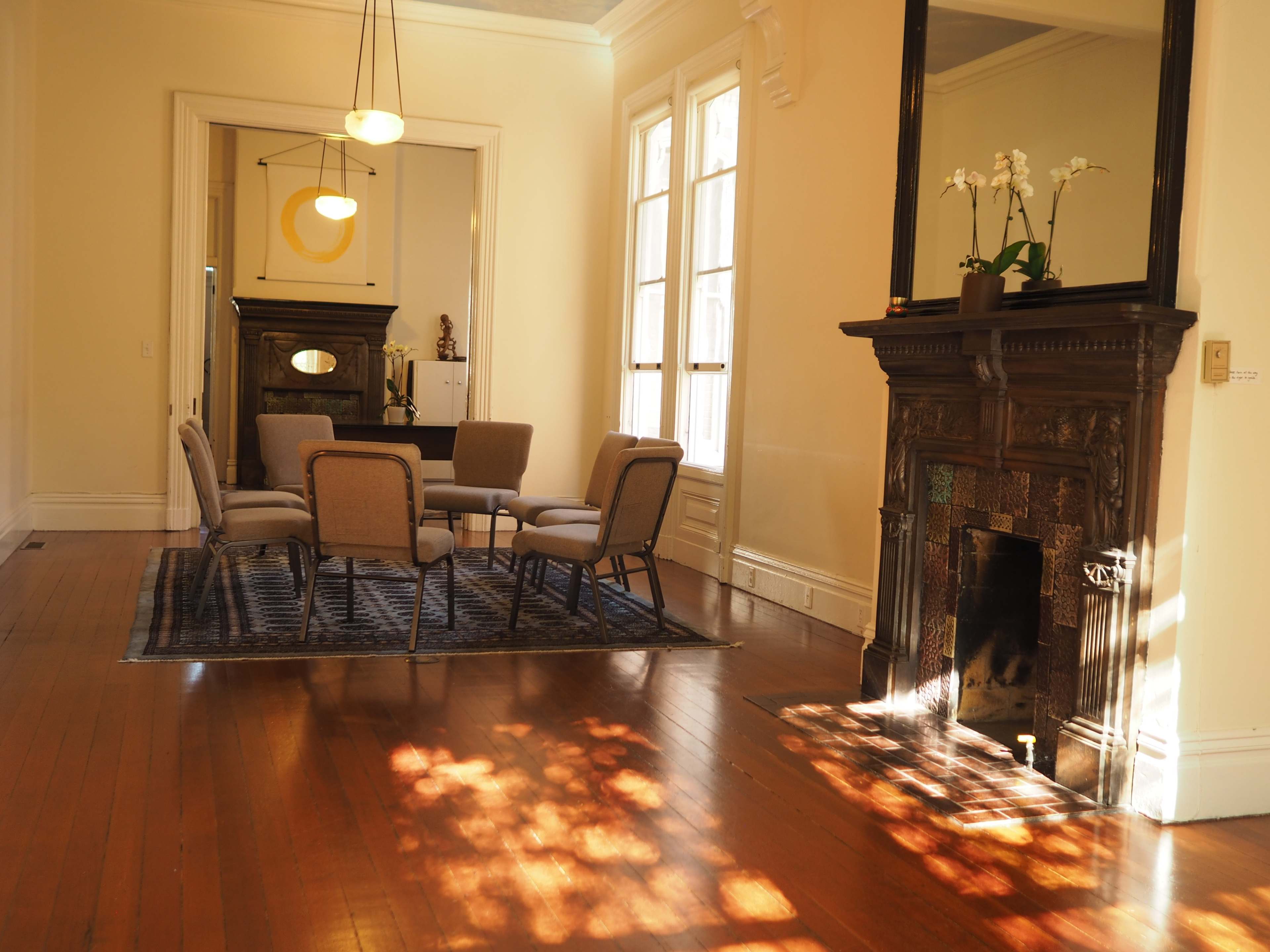 The image shows a spacious room with a circular arrangement of chairs on a patterned rug, a decorative fireplace, and large windows that allow natural light to enter.