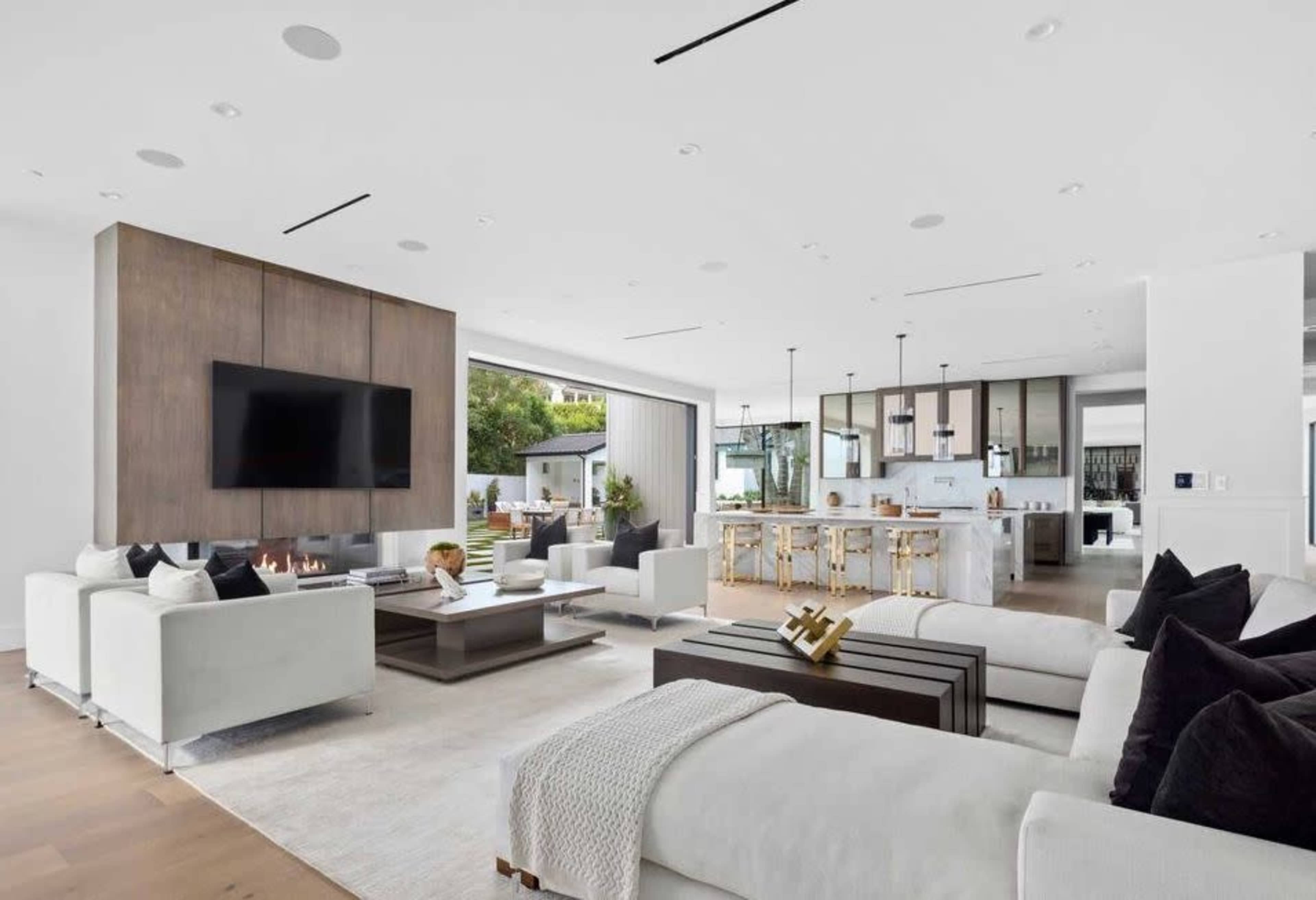 A modern living room features a large flat-screen TV mounted on a wooden panel, with white sofas arranged around a central coffee table and an open kitchen visible in the background.