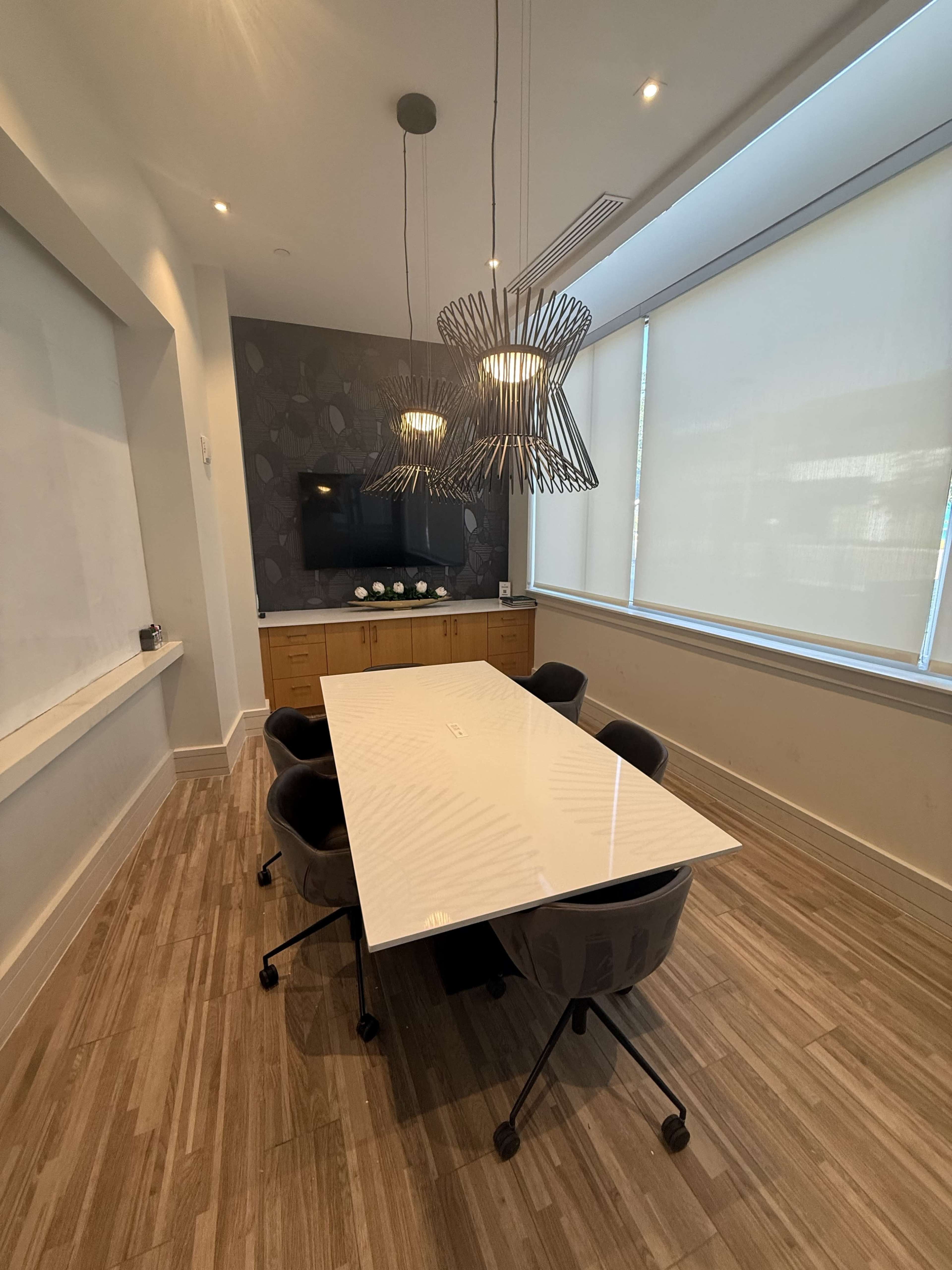 The image shows a modern conference room featuring a large white table, four gray chairs, and two decorative light fixtures hanging from the ceiling.
