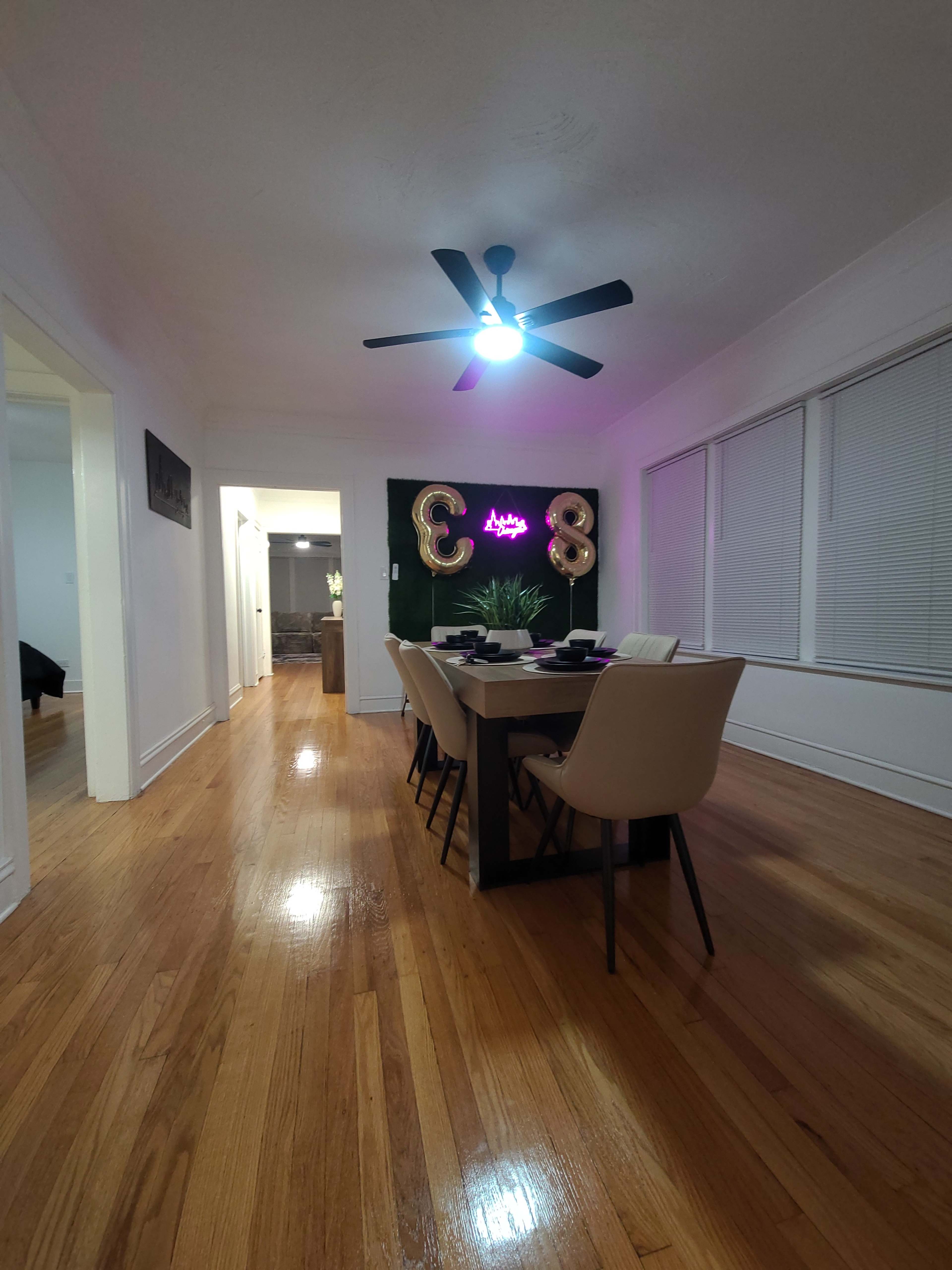 A dining room features a large table set for a meal, with decorative balloons and a vibrant wall backdrop illuminated by a ceiling fan light.