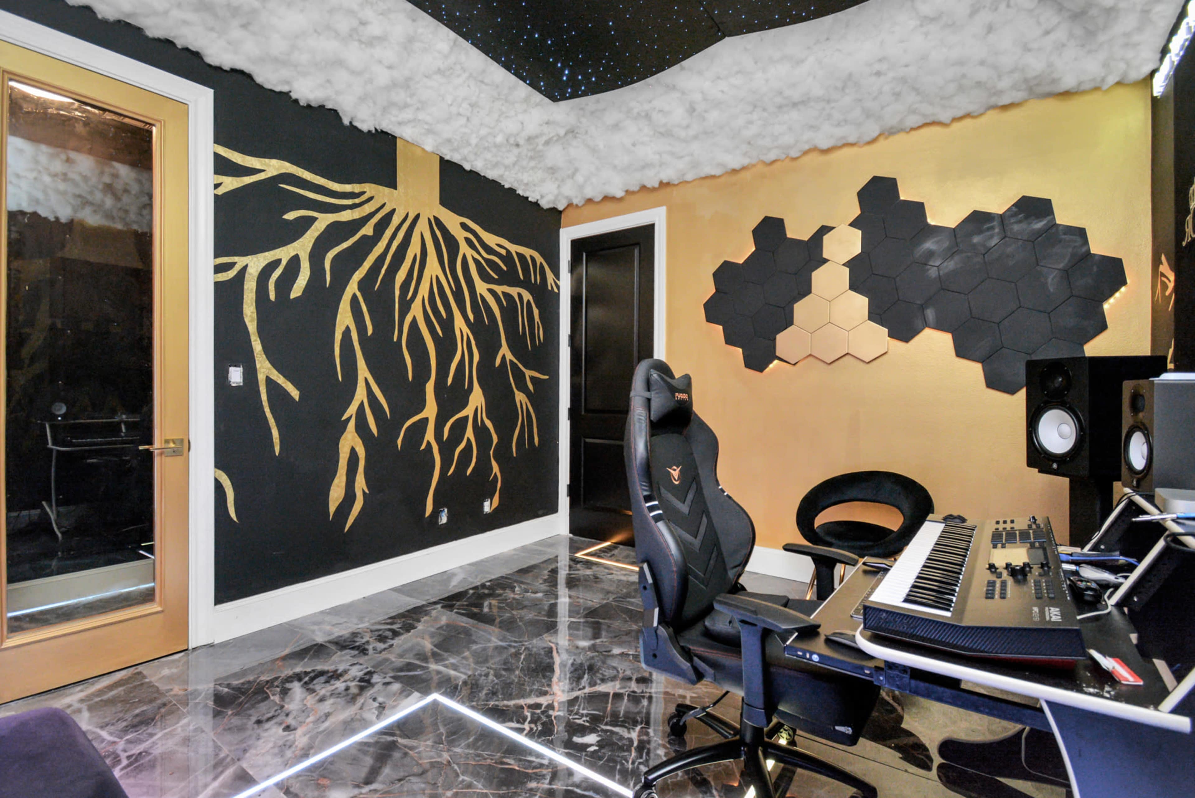 The image shows a modern music studio featuring a black and gold color scheme, a large black desk with a keyboard and studio monitors, and unique wall art.