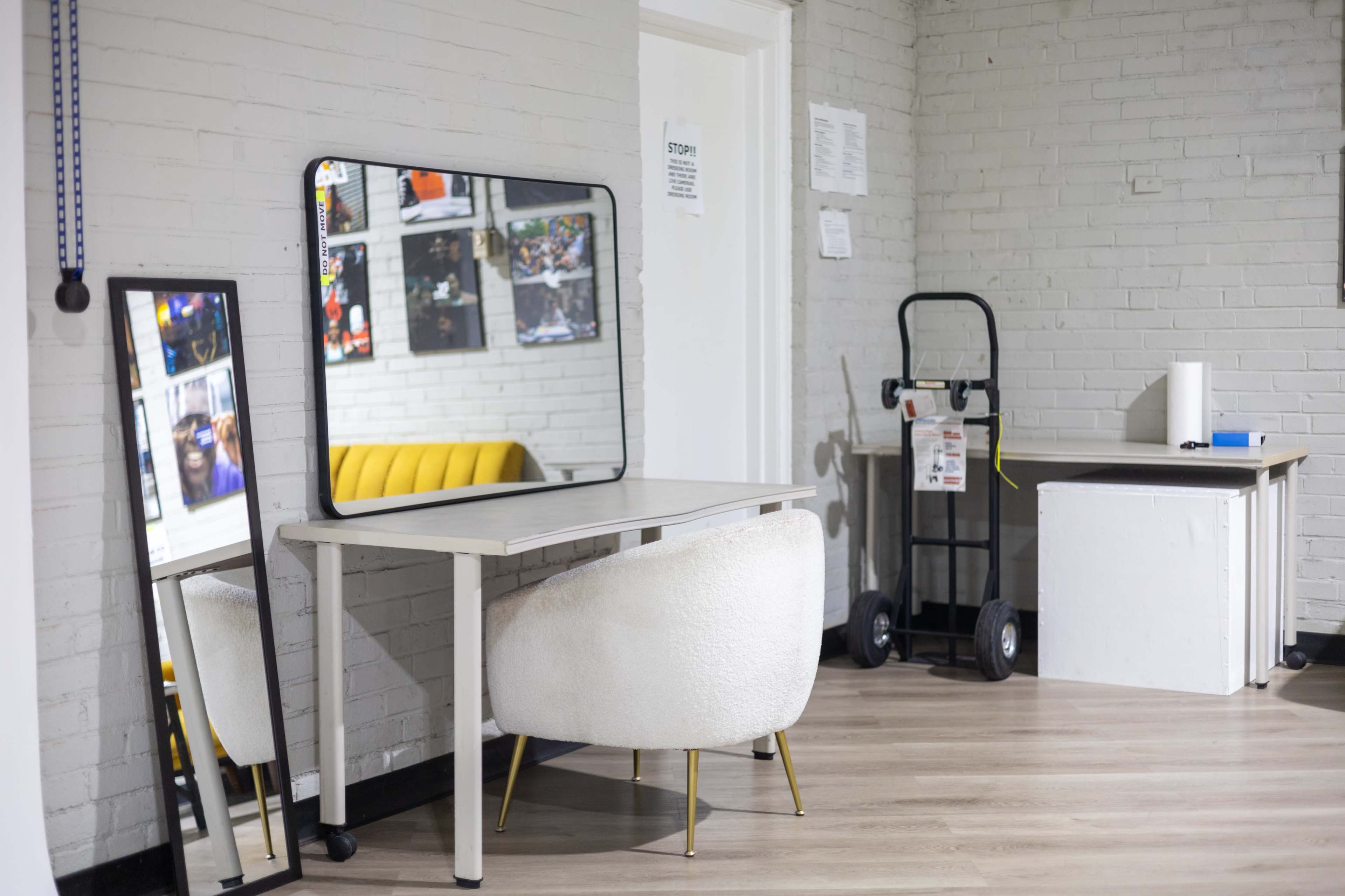 The image shows a bright, modern room featuring a white desk with a large mirror, a stylish chair, and a utility cart in the background.