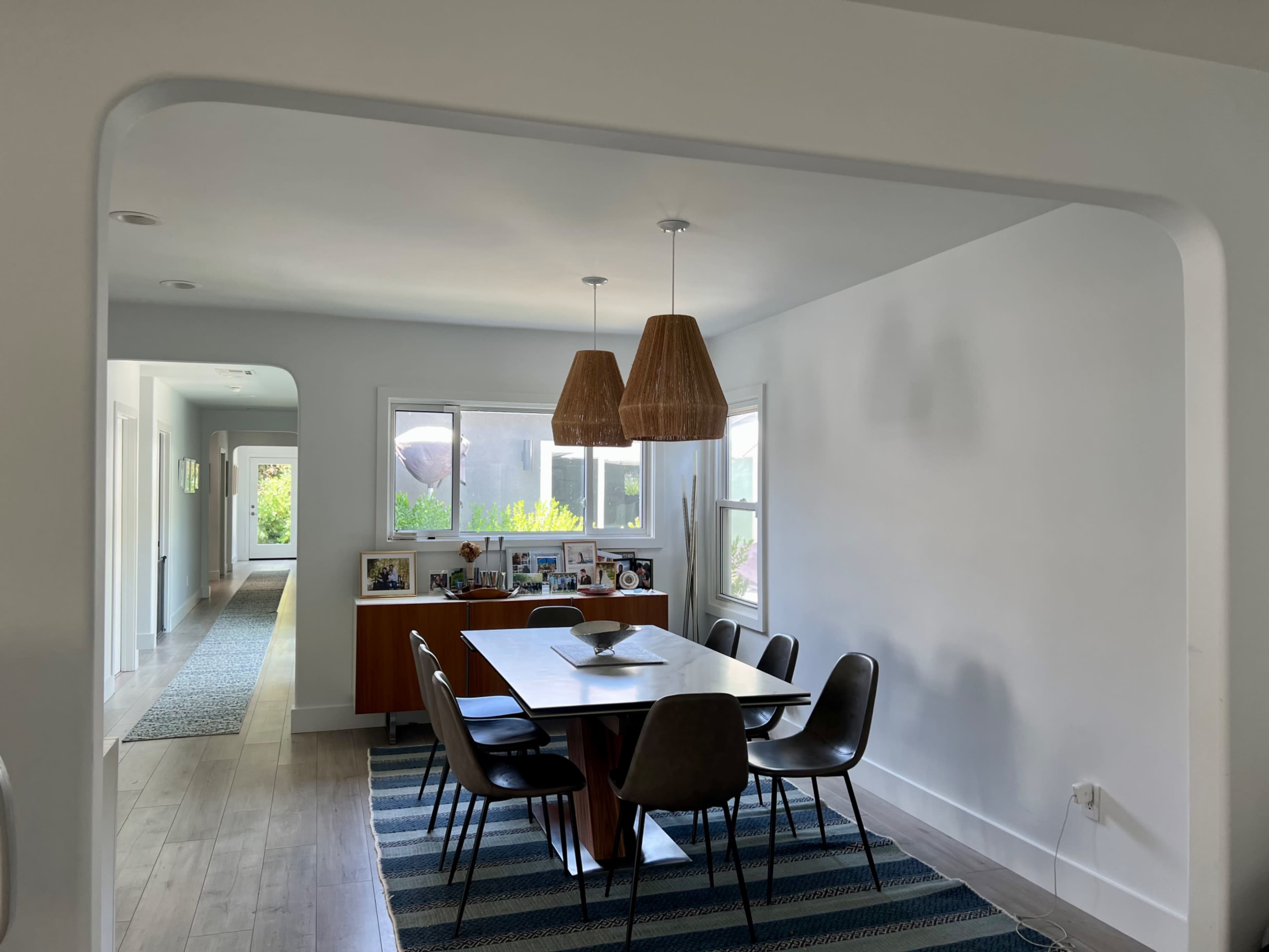 The image shows a dining area with a table surrounded by chairs, two pendant lights hanging above, and a view of a hallway through an archway.