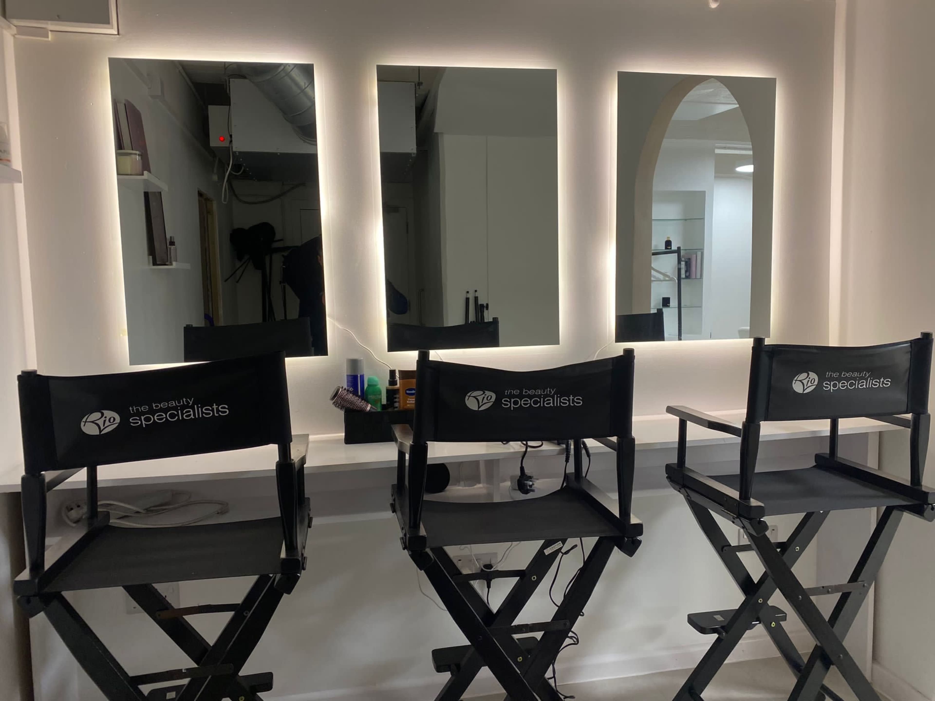 Three director's chairs are arranged in front of three illuminated mirrors in a beauty salon.