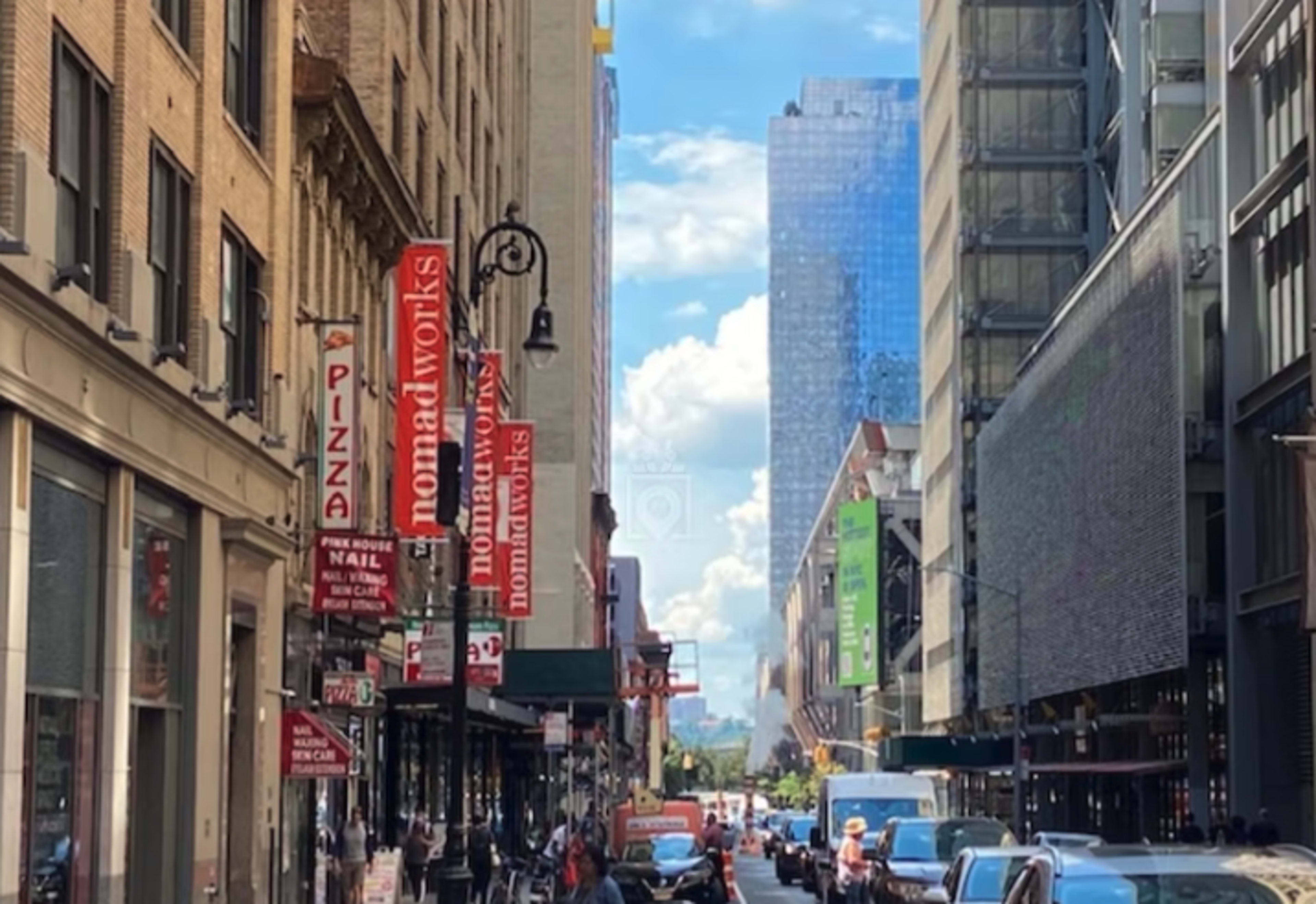 A bustling city street features storefronts with signs for pizza, nail salons, and a modern glass building against a backdrop of clouds.