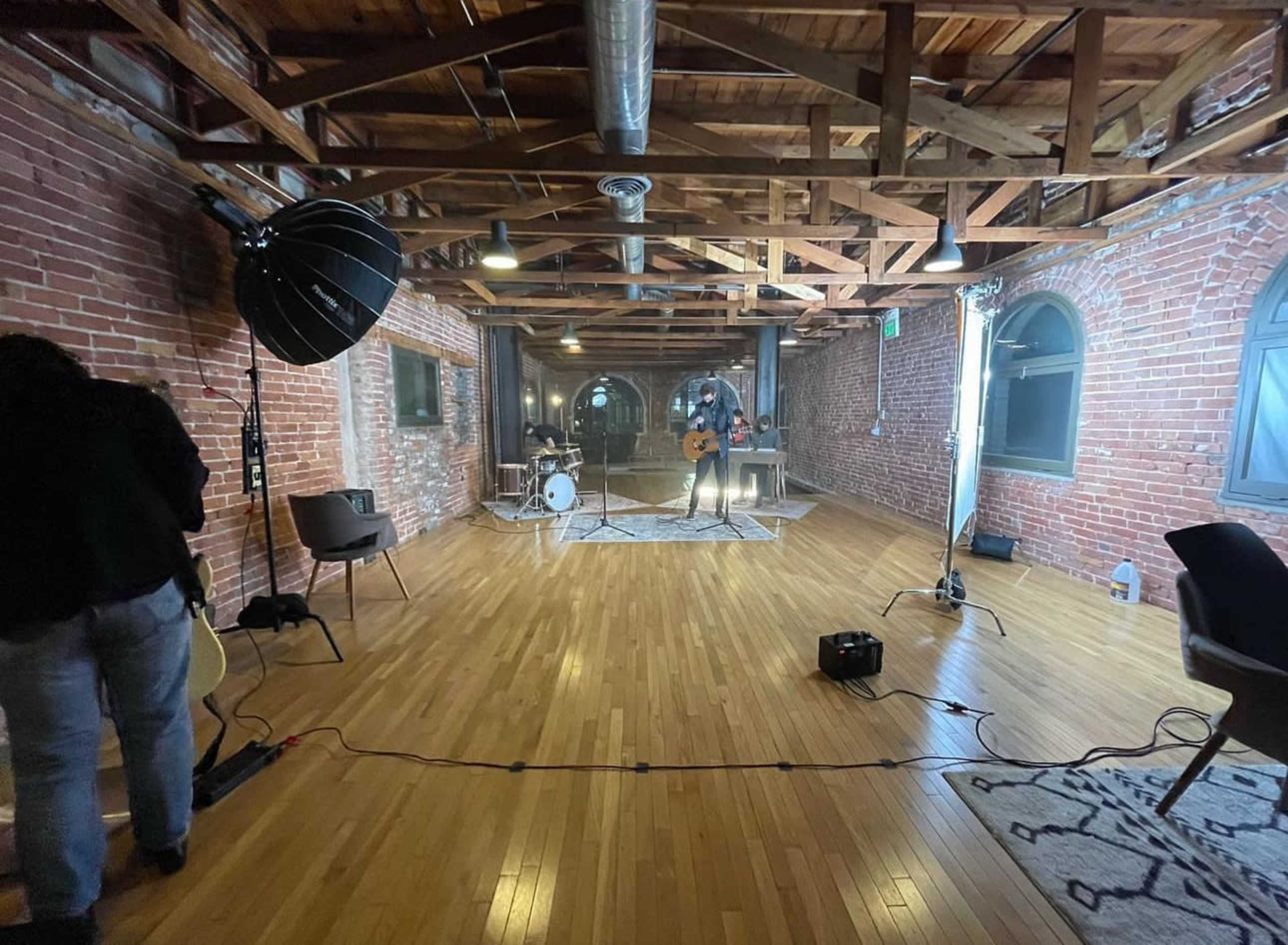 A musician is performing with a guitar in an empty, well-lit room featuring exposed brick walls and wooden beams.