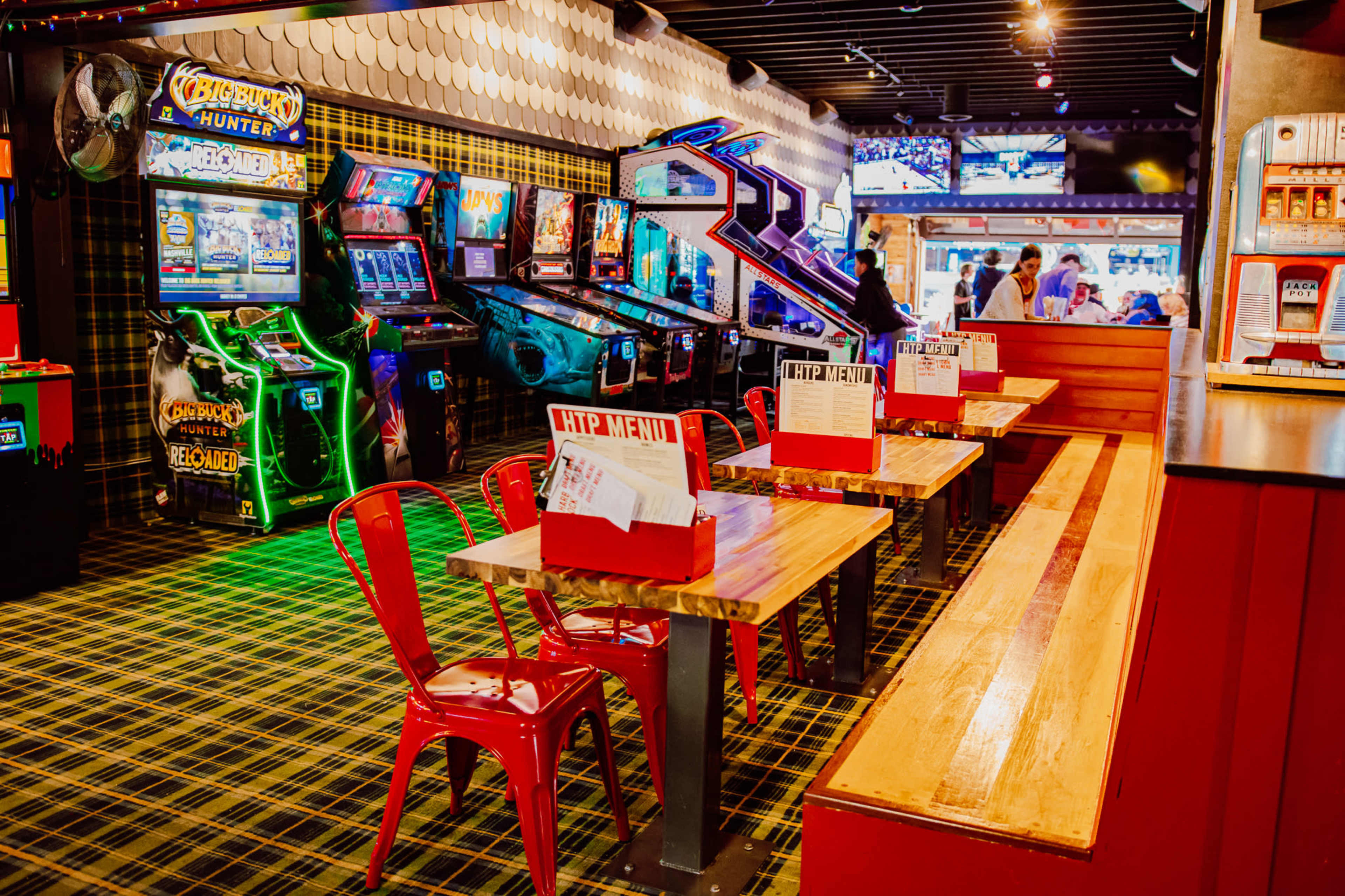 The image shows a lively game arcade with brightly lit machines and empty tables featuring red chairs against a checkered floor.