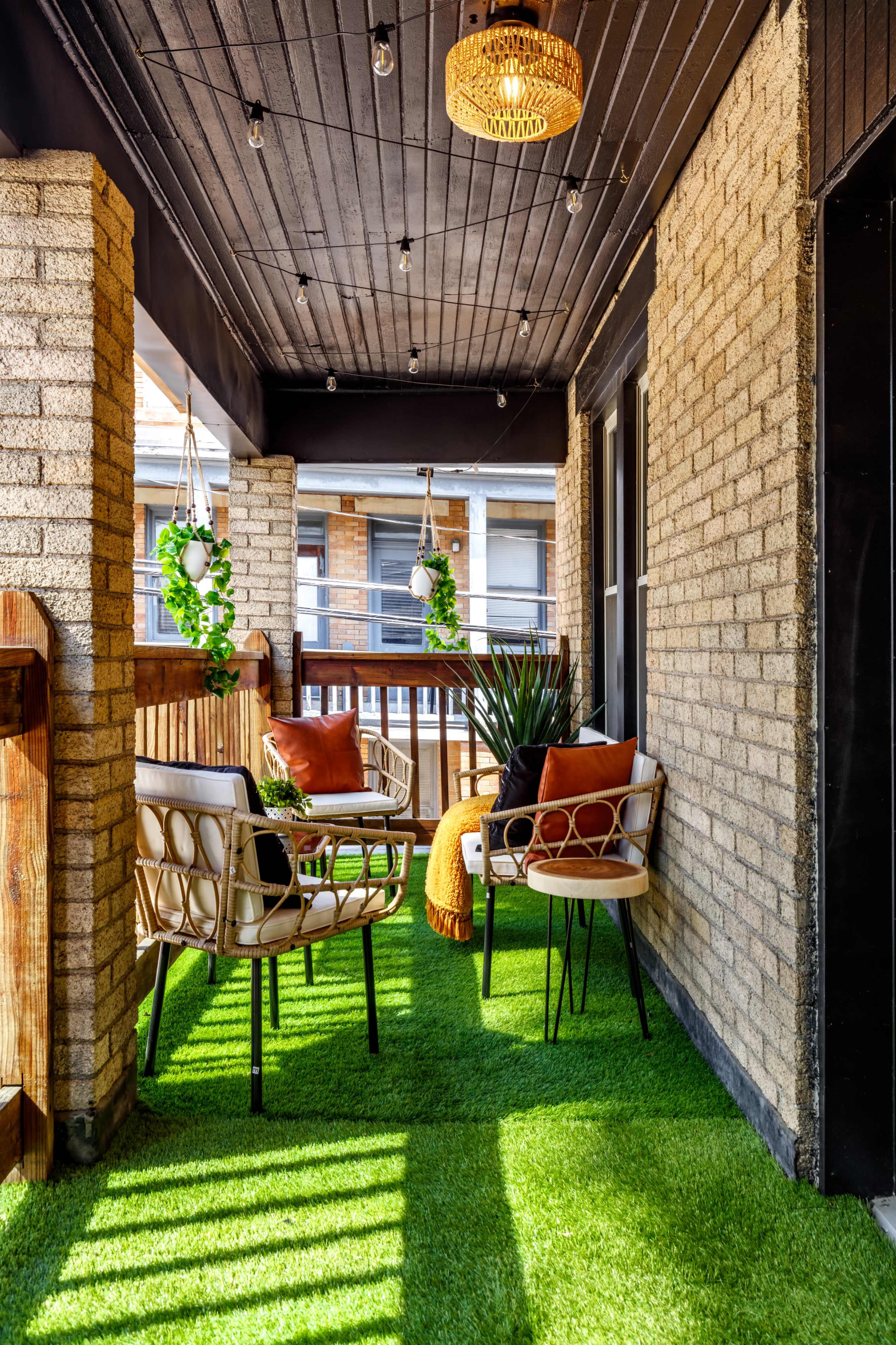 The image shows a balcony featuring two chairs, a small table, potted plants, and decorative lights, all set on artificial grass.