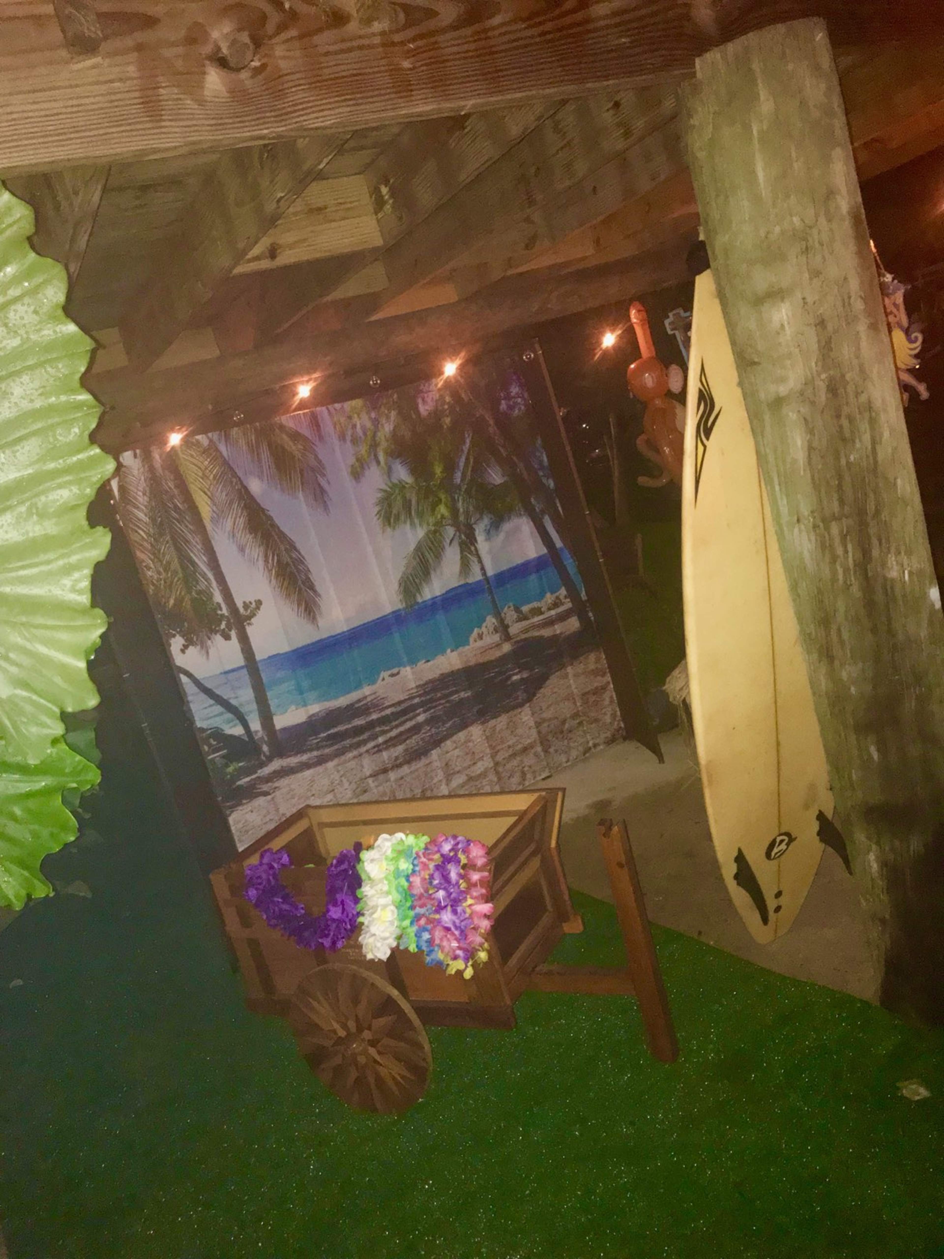 The image shows a decorated outdoor space with a surfboard, a cart holding colorful leis, and a backdrop of a tropical beach scene.