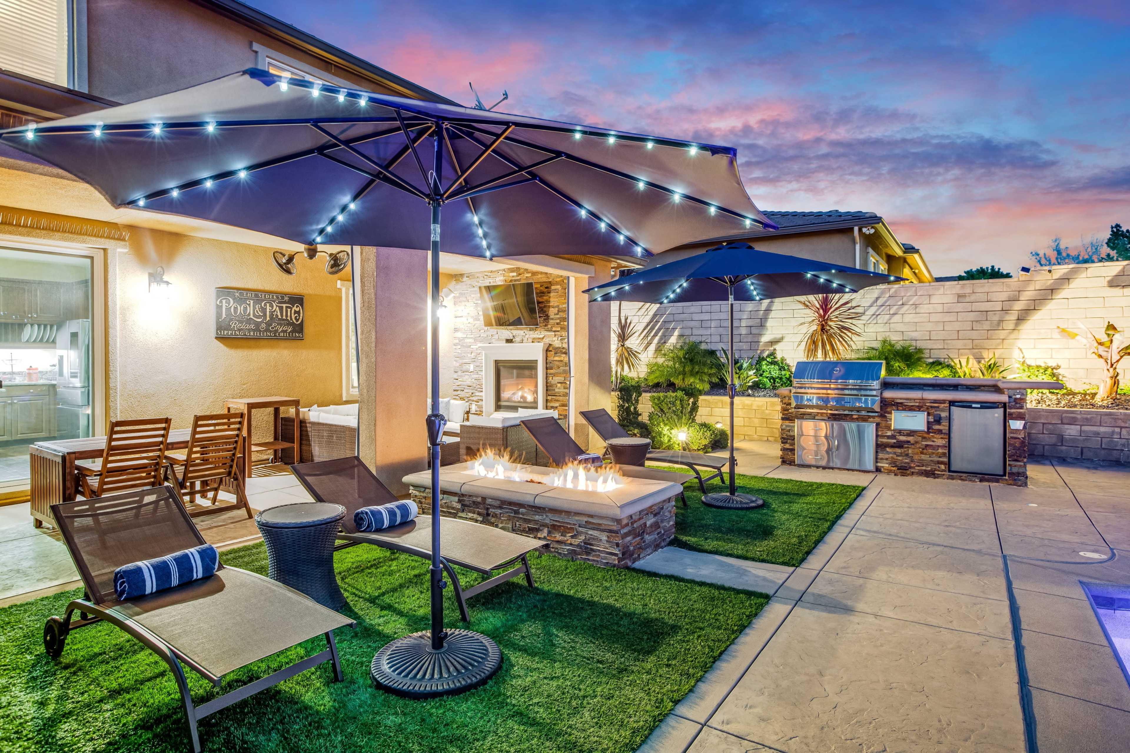 The image shows a backyard patio featuring two large umbrellas, lounge chairs, a fire pit, and an outdoor kitchen, set against a colorful sunset sky.
