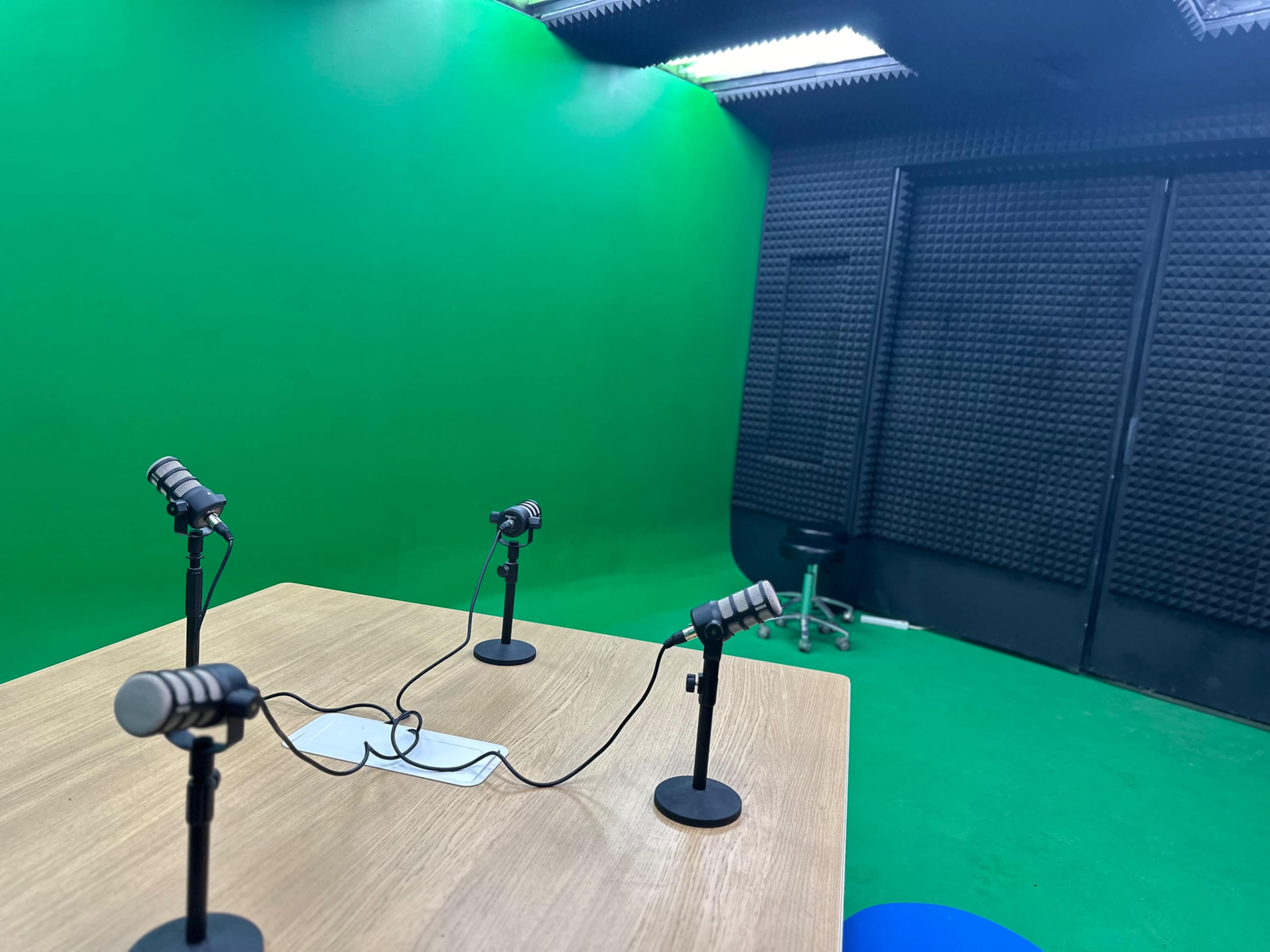 The image shows a podcast recording studio featuring a table with microphones and a green screen background.