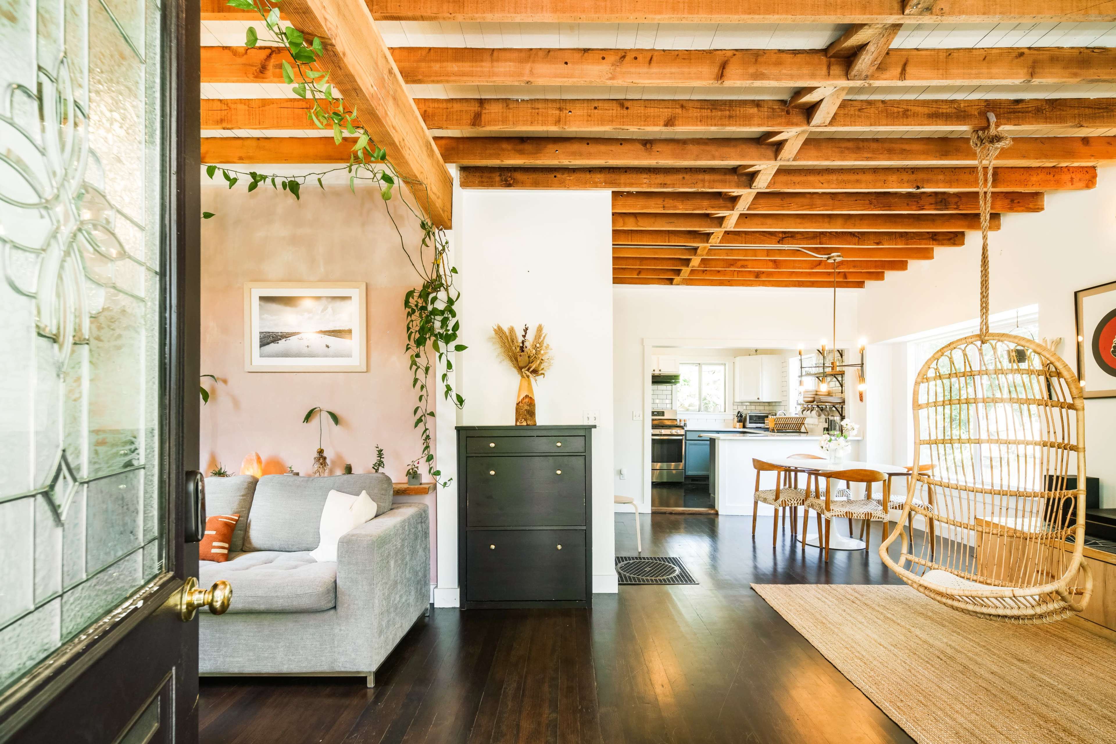 A cozy living space features exposed wooden beams, a hanging chair, and a dining area adjacent to a bright kitchen.