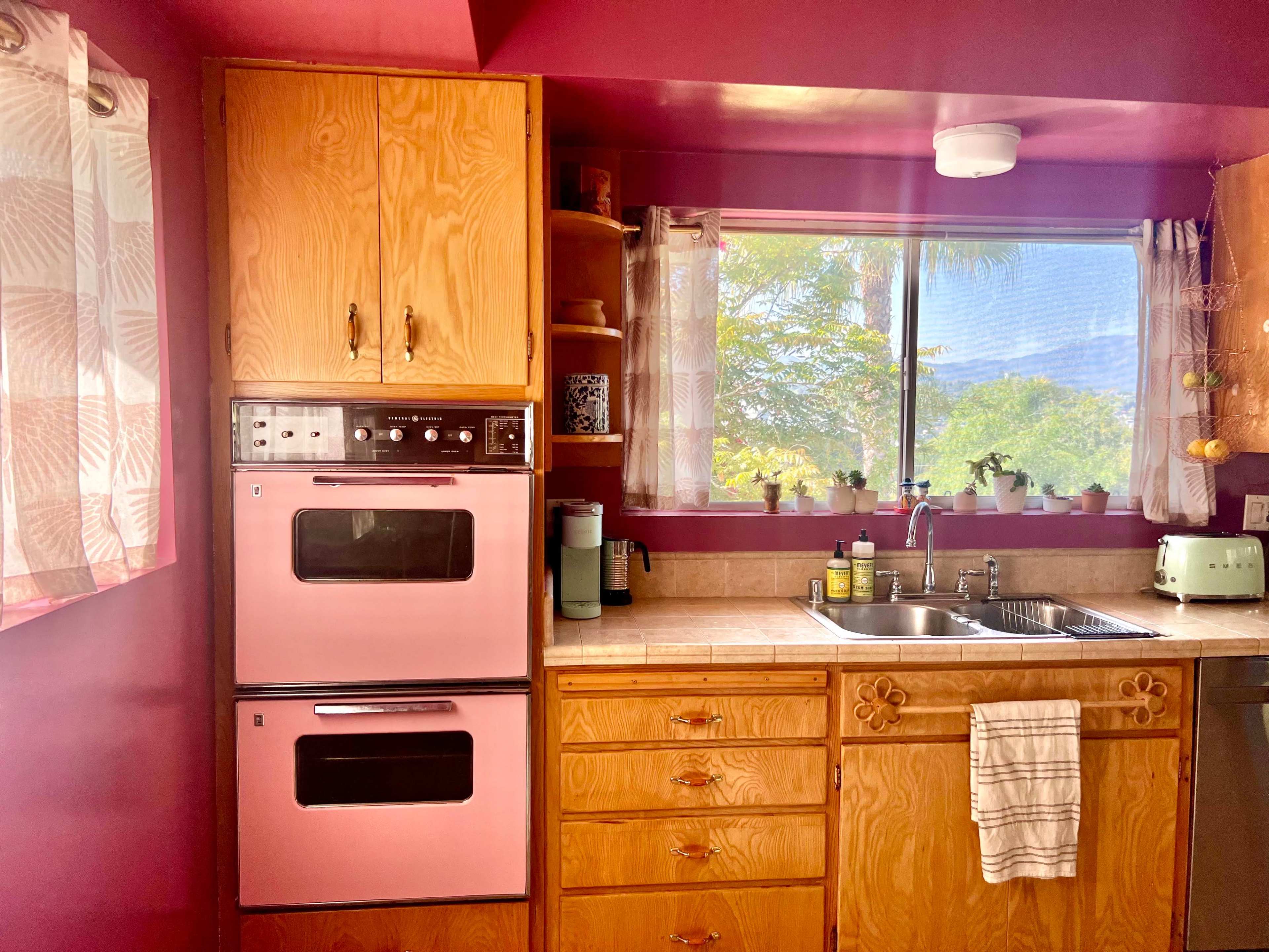 The image shows a kitchen with pink walls, wooden cabinets, and a window featuring a view of greenery outside.