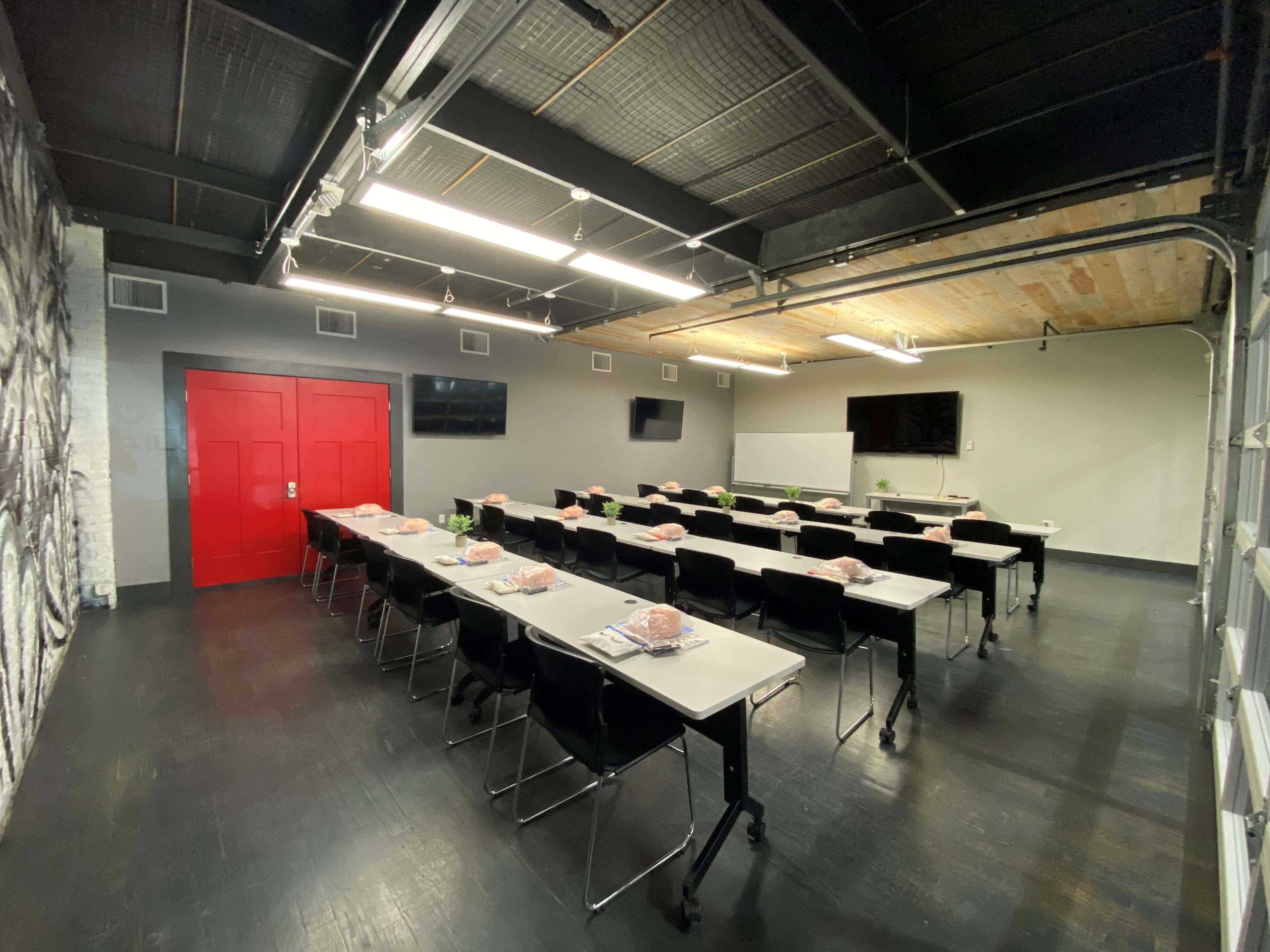The image features a modern classroom setup with rows of tables and chairs, two large screens on the walls, and a bold red door.