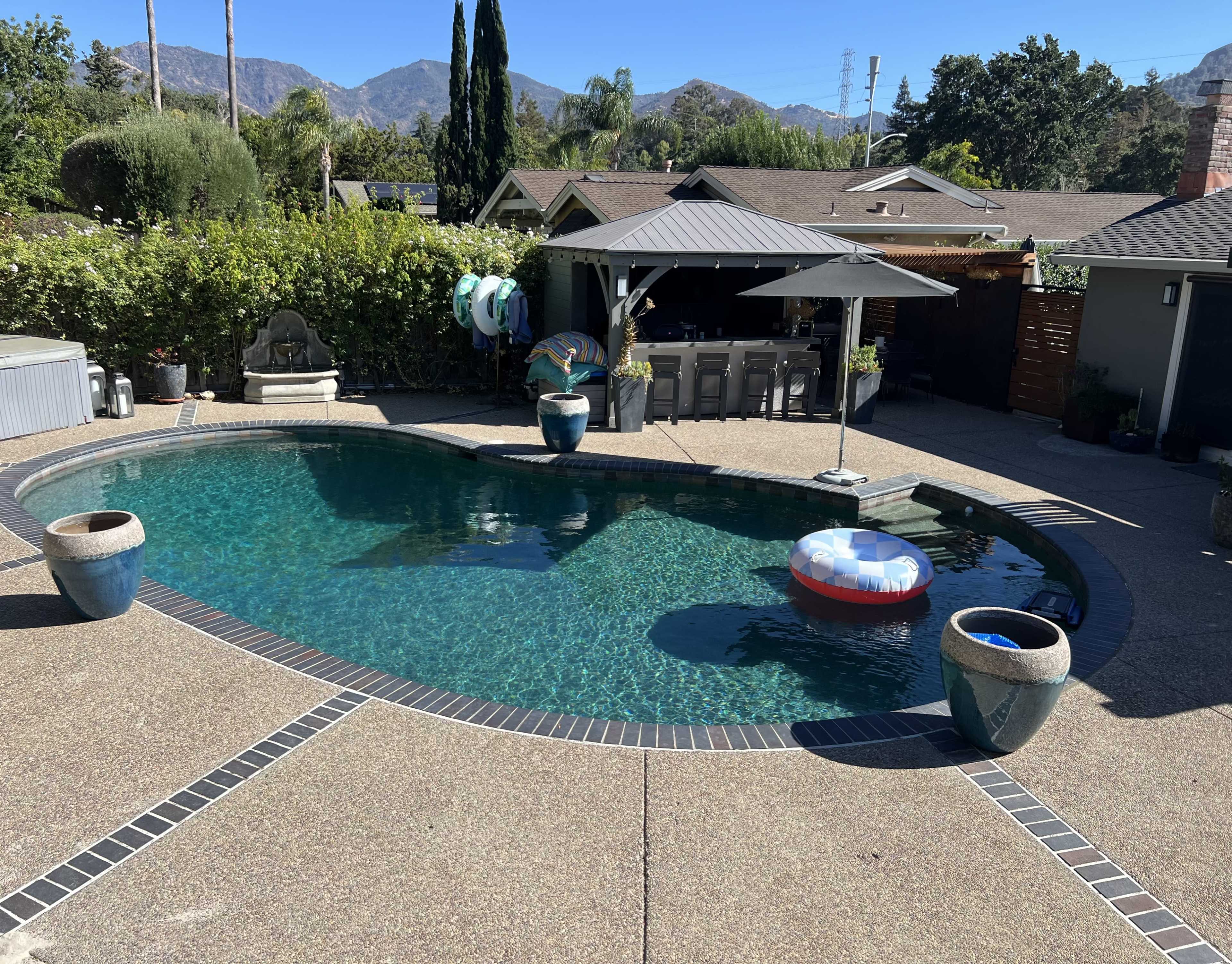The image shows a backyard with a swimming pool, surrounded by patio areas, decorative plants, and a covered outdoor seating space.