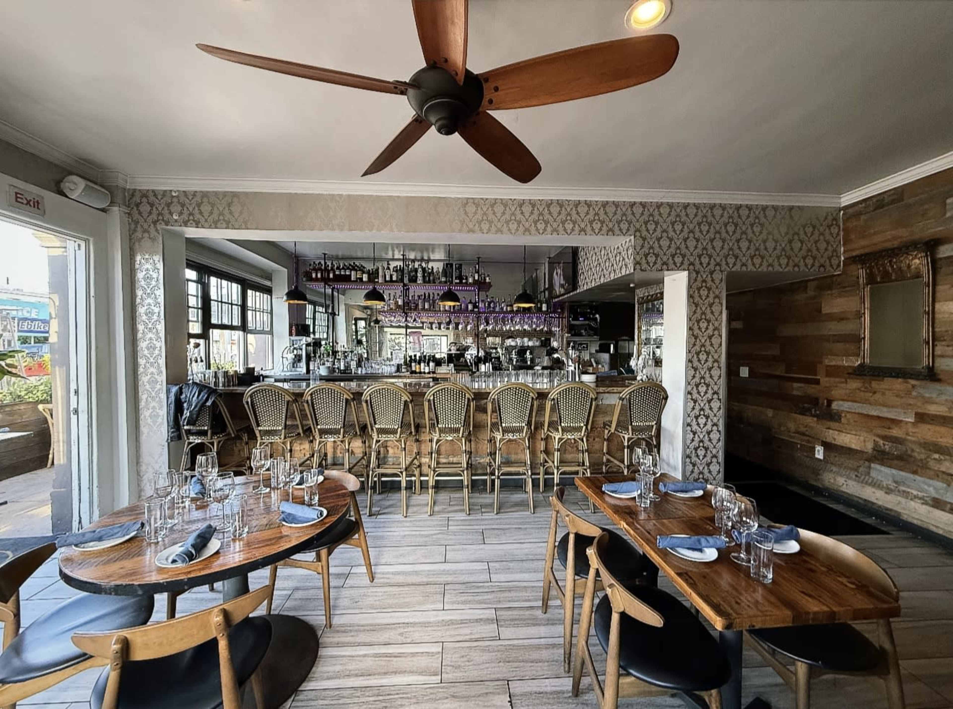 The image shows a restaurant interior with wooden tables and a bar lined with high chairs, featuring bottles displayed on shelves behind the bar.