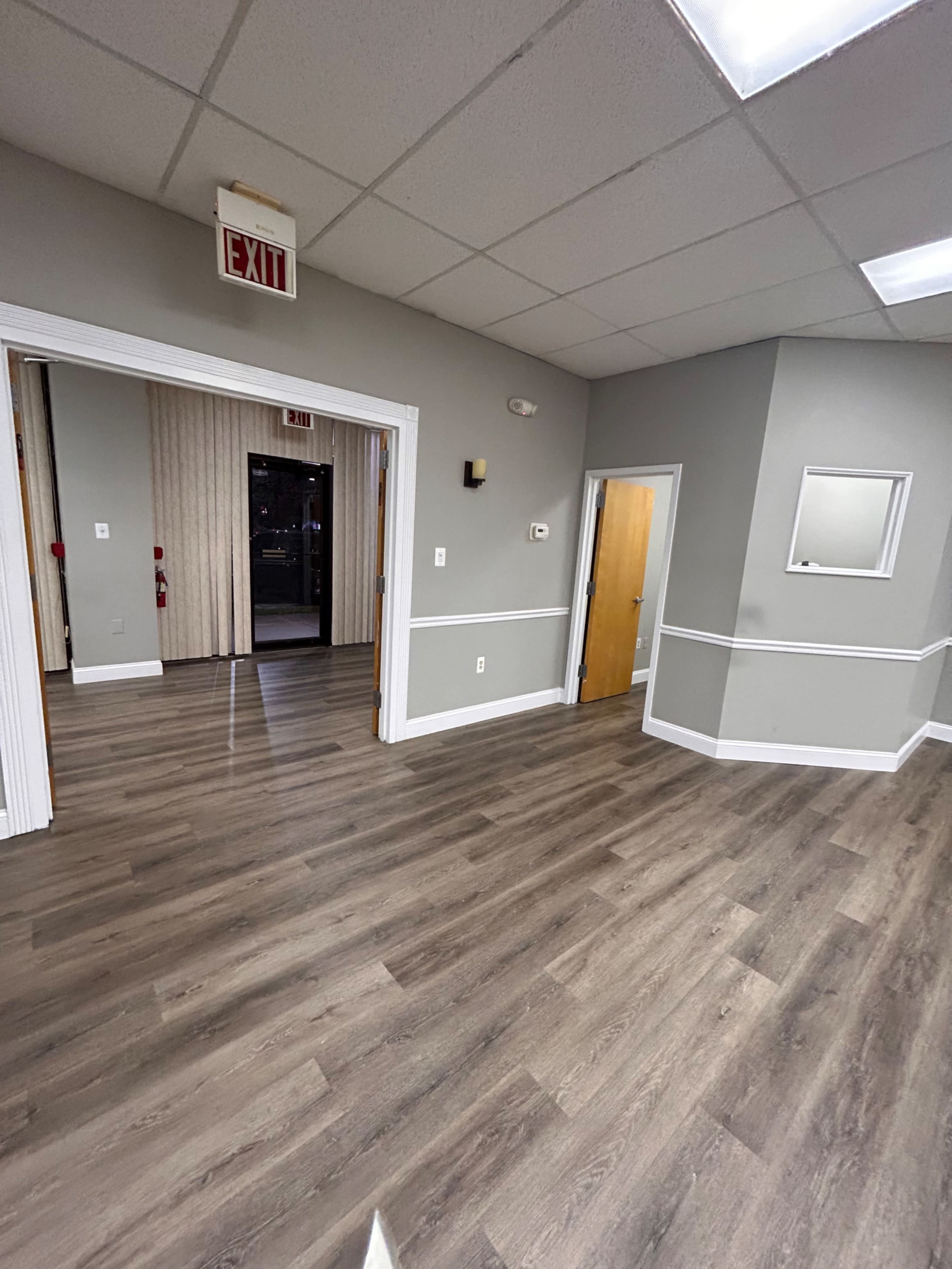 A spacious, empty room with gray walls and wooden flooring, featuring an exit sign and a doorway leading to another area.