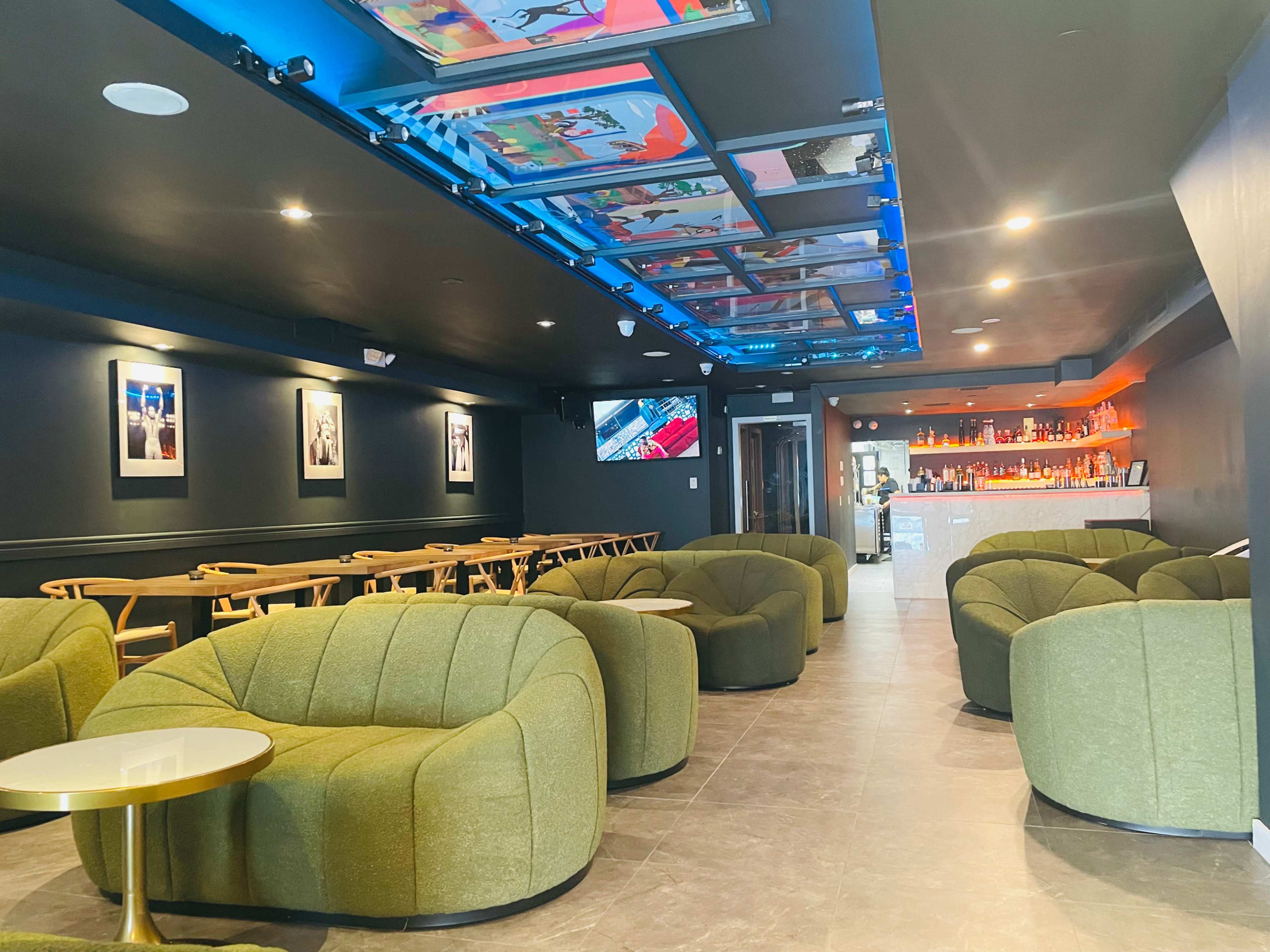 A modern lounge area with green upholstered chairs, a bar in the background, and colorful artwork displayed on the ceiling.