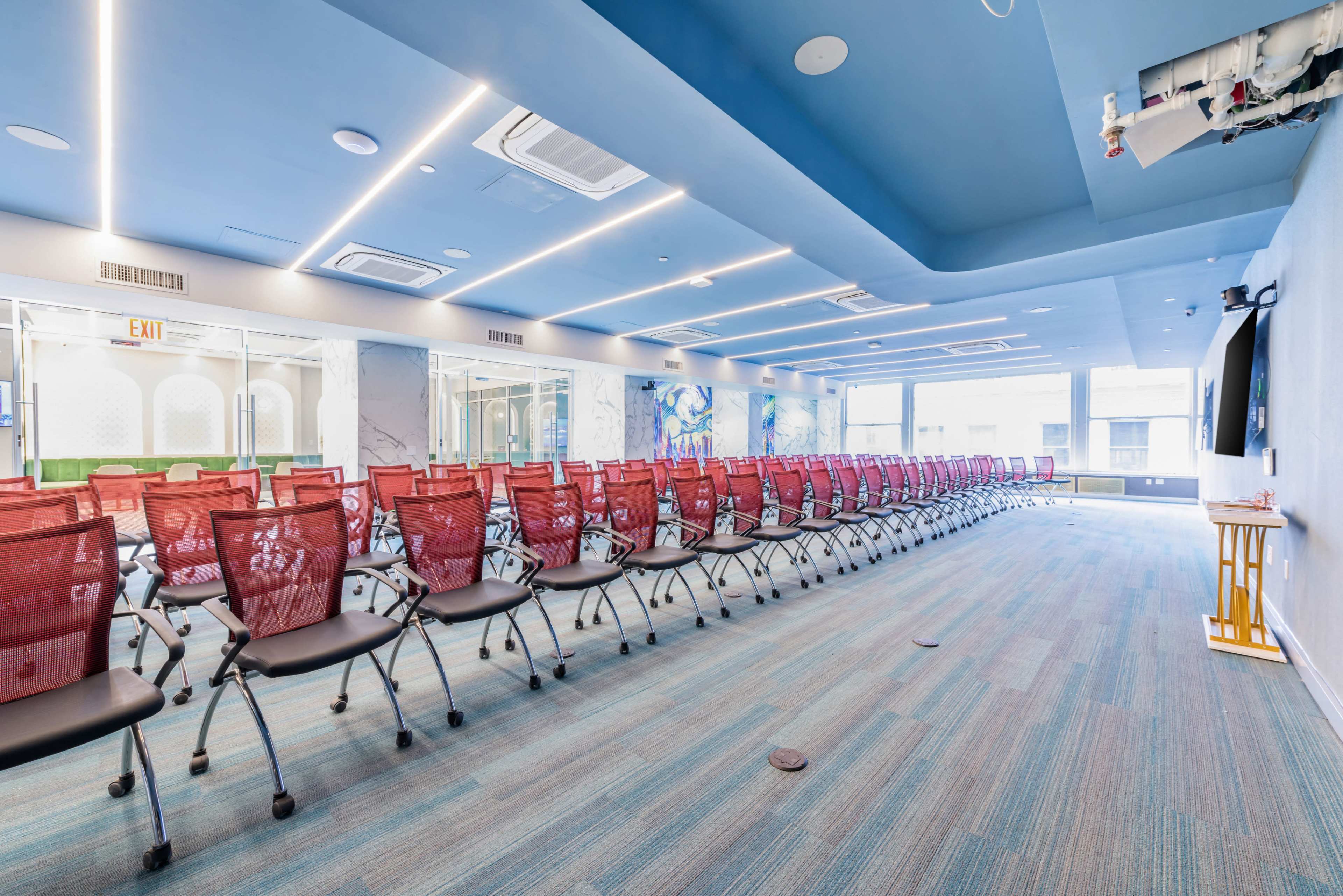 The image shows a modern conference room with rows of red chairs arranged facing a screen, and large windows allowing natural light to enter.