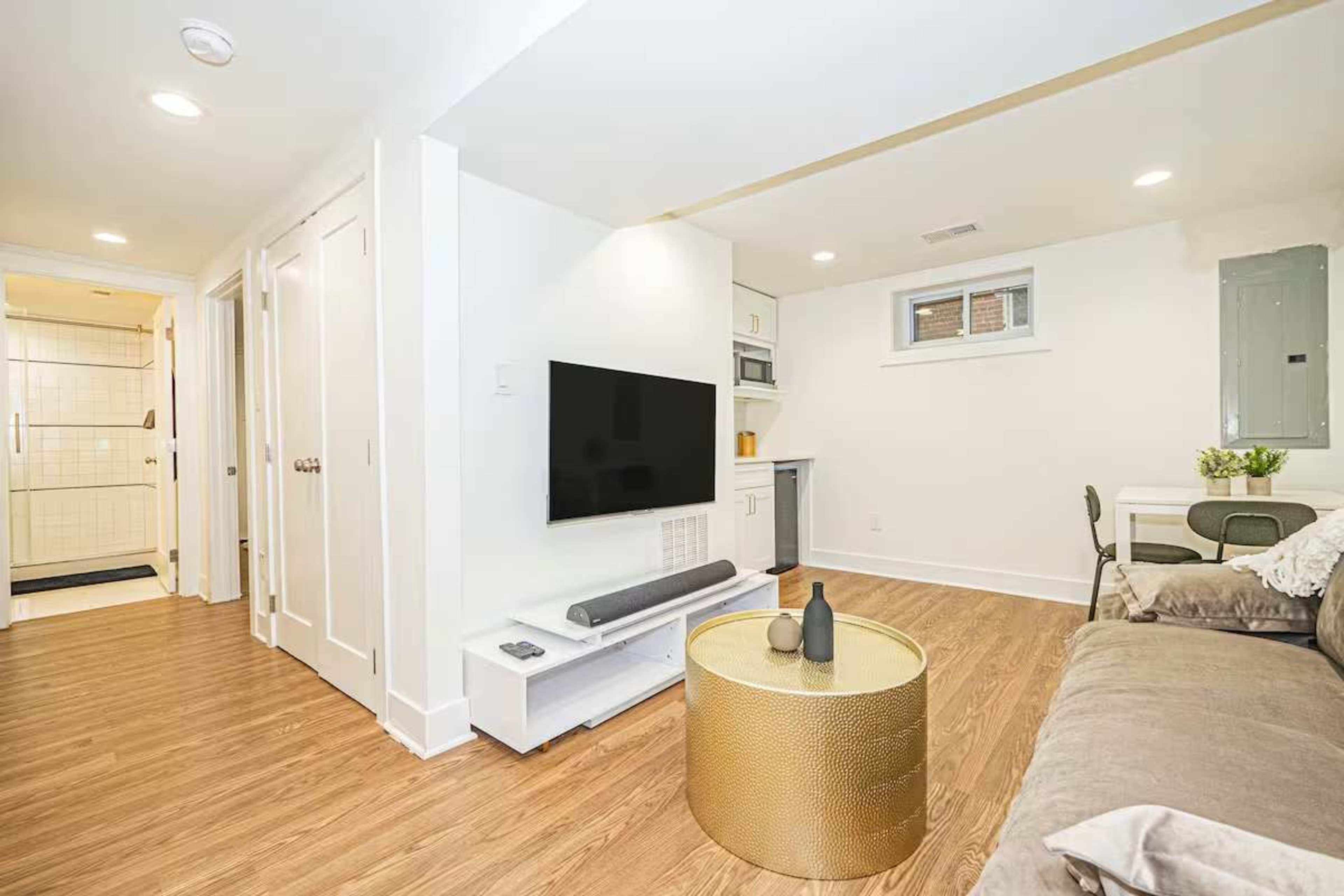 The image shows a modern, minimalist living space with a television mounted on the wall, a brown couch, and a small dining area in the corner.