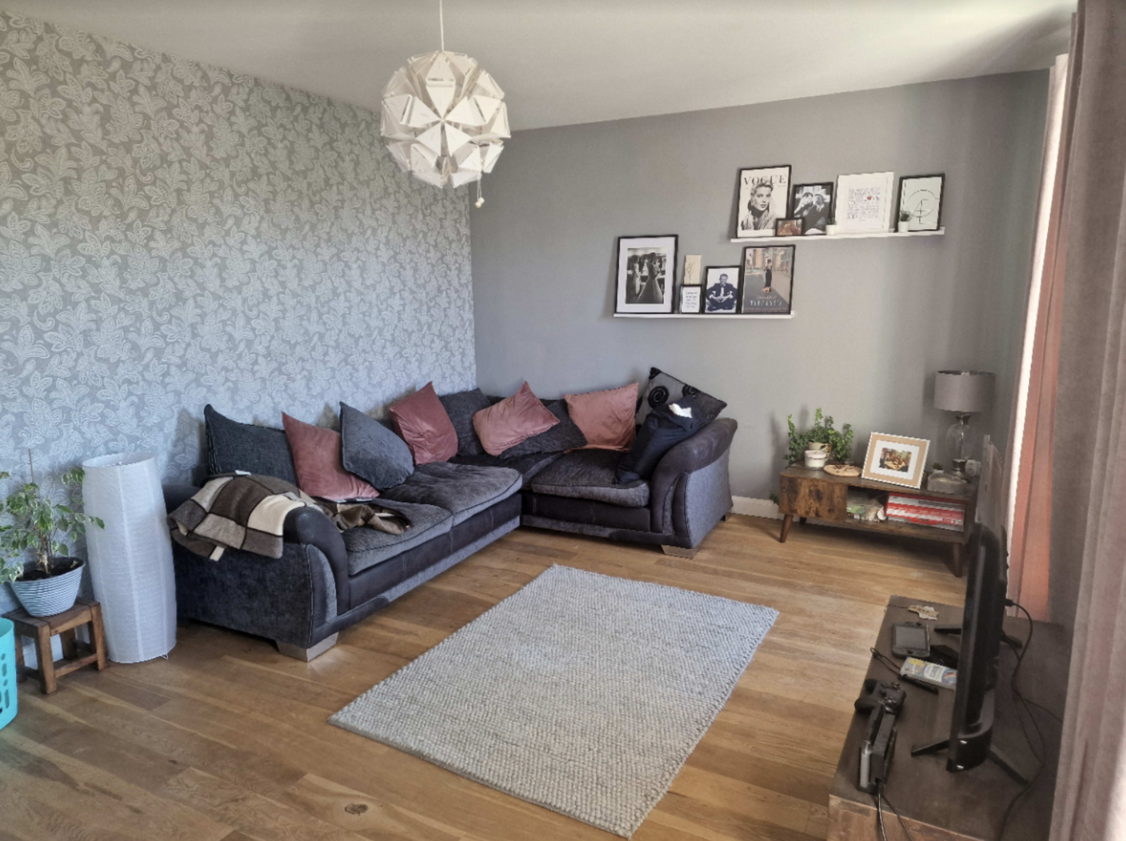 A living room with a dark sectional sofa, decorative cushions, a light rug on wooden flooring, and framed artwork on the wall.