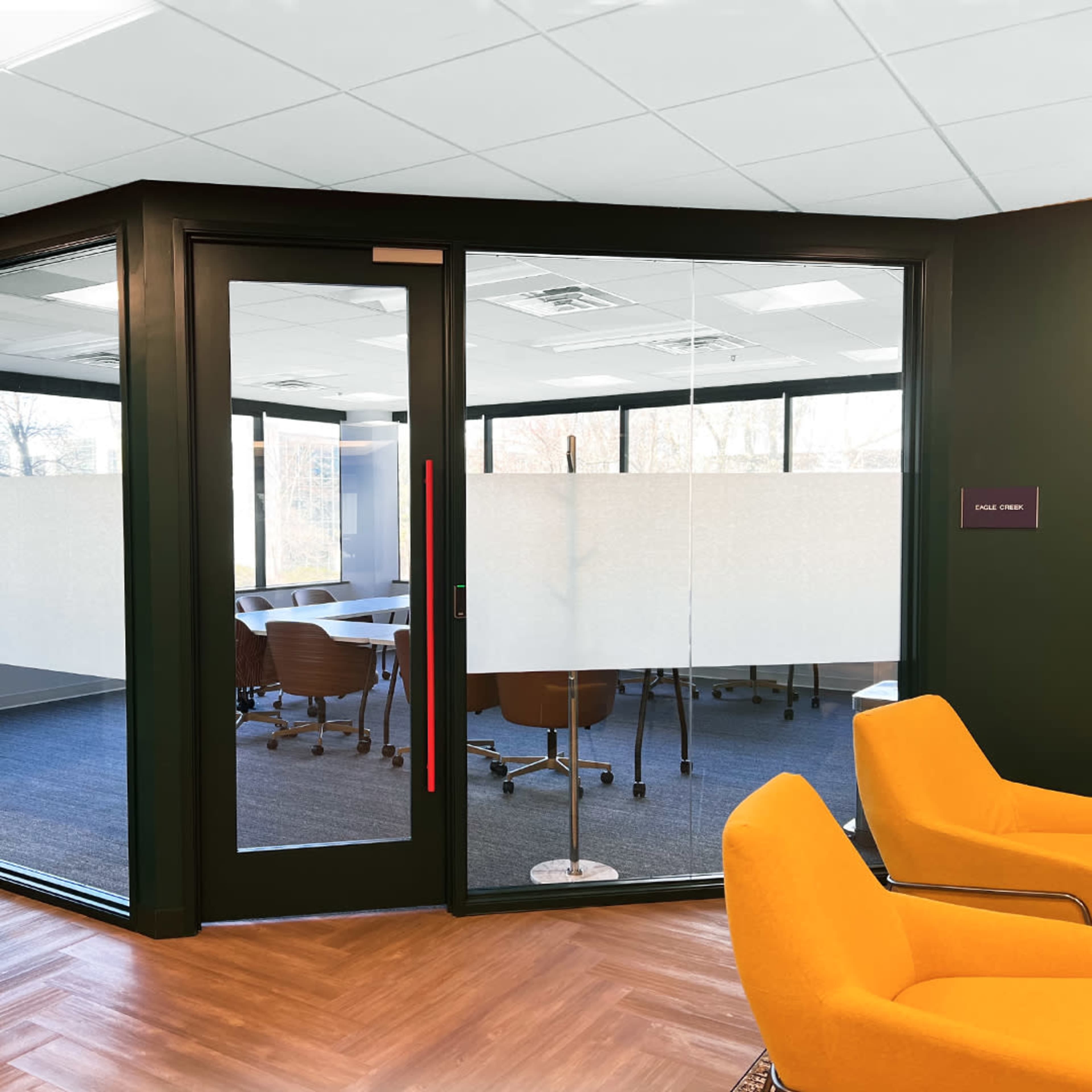 A modern conference room with glass doors, featuring a large table surrounded by brown chairs and a view of a bright, open workspace.