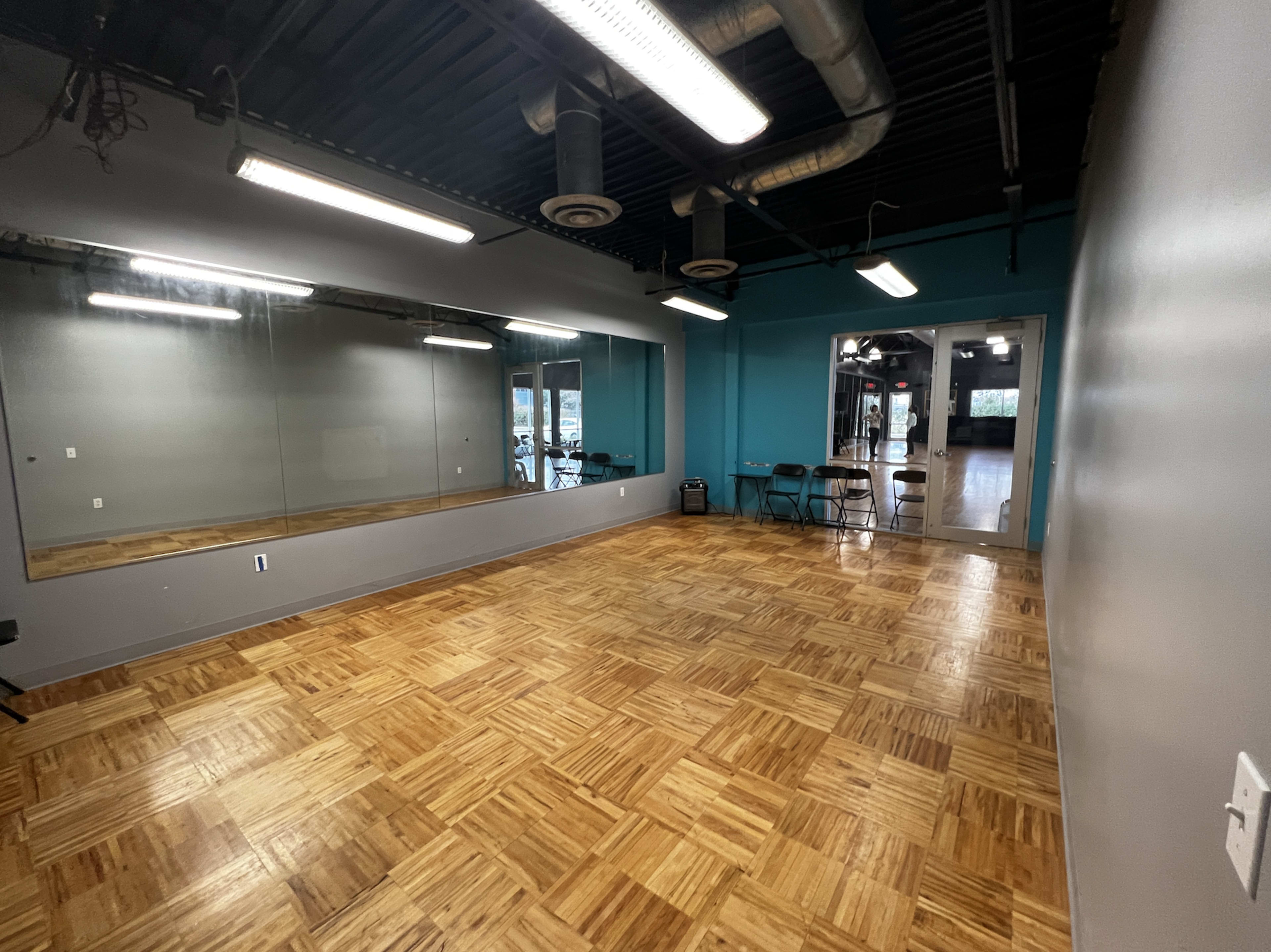 The image shows a spacious room with a wooden floor, mirrors on one wall, and teal-colored accent walls.