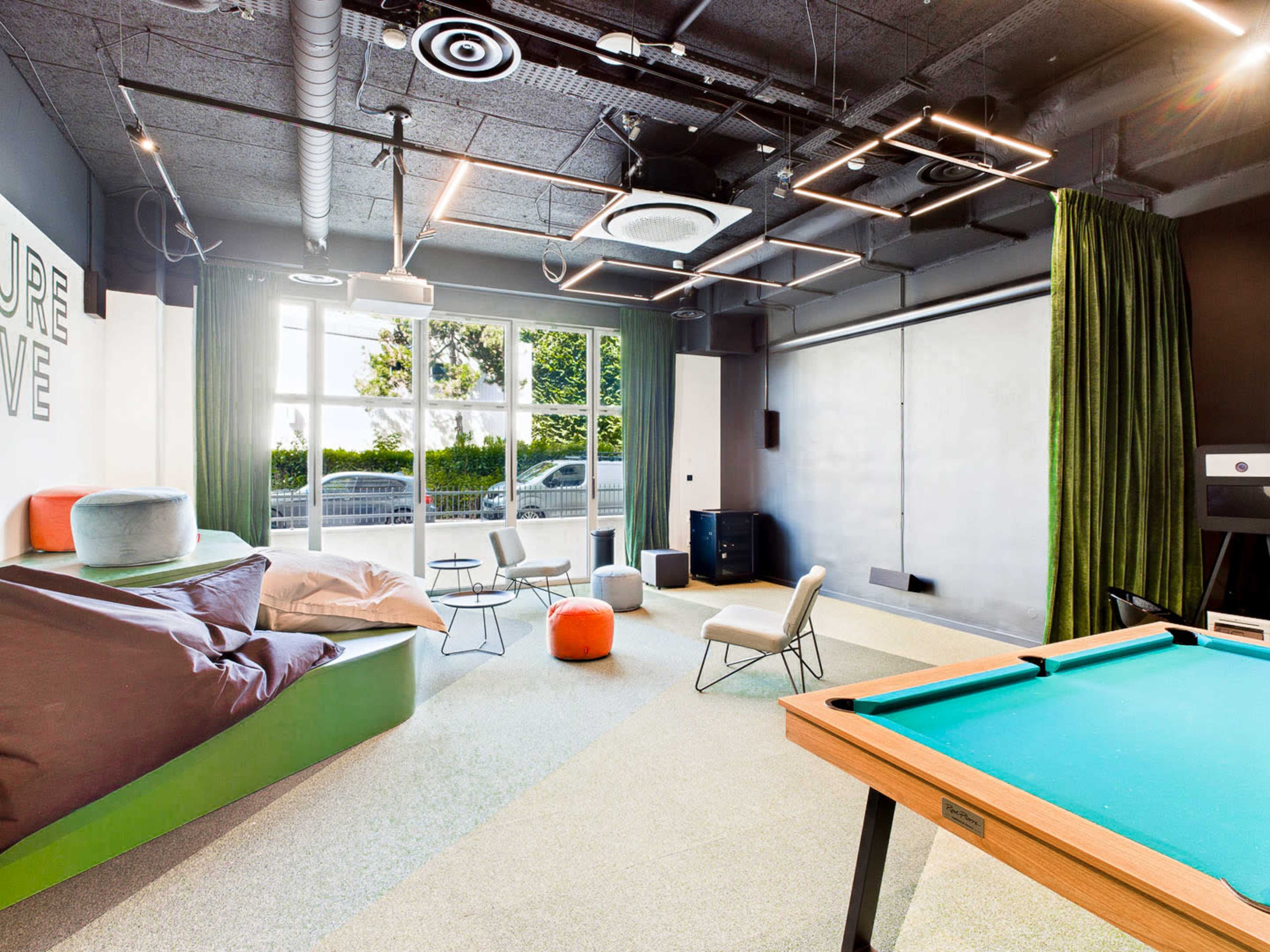The scene shows a modern recreational room featuring a billiard table, bean bags, and comfortable seating, with large windows letting in natural light.