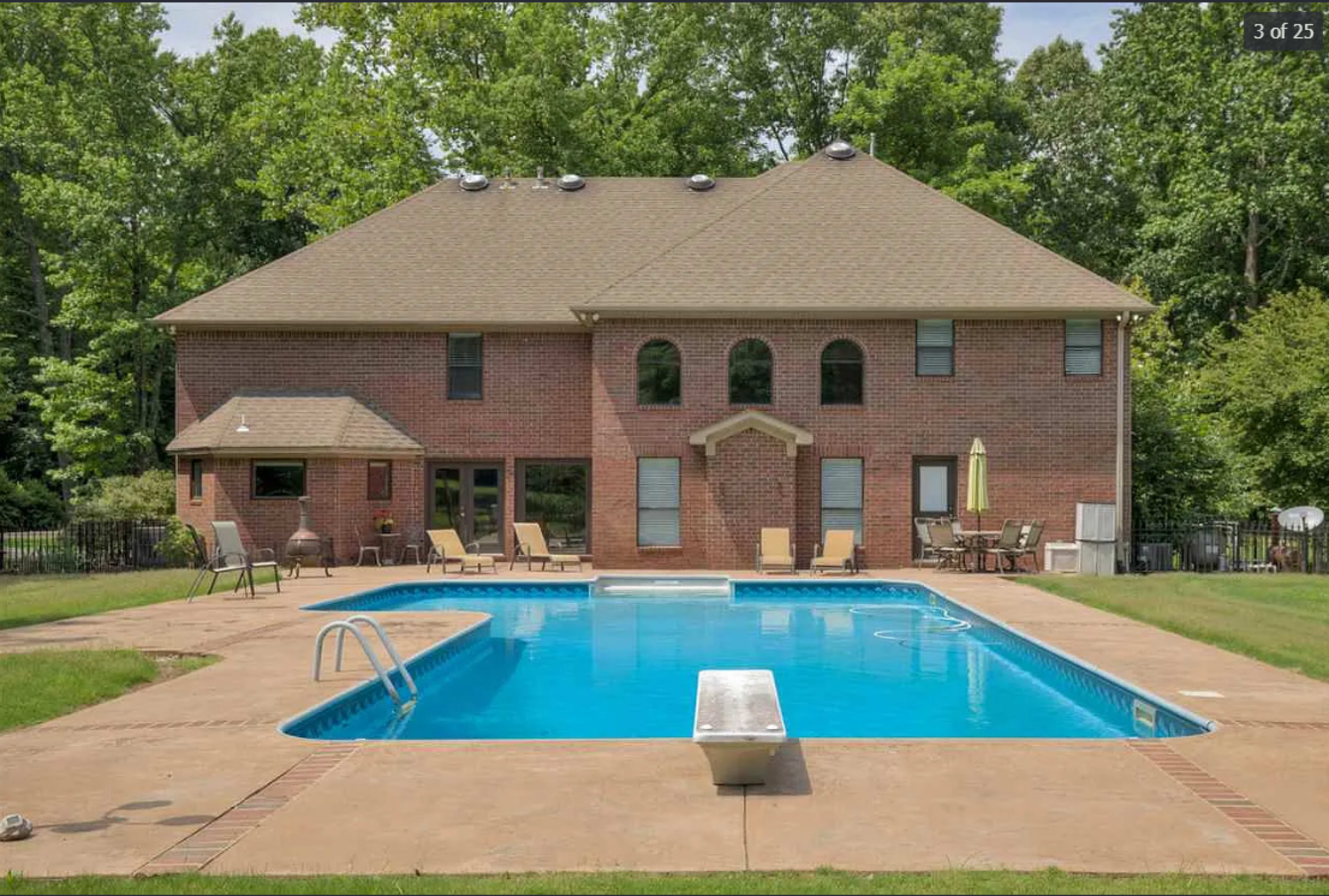 The image shows a two-story brick house with multiple windows and a spacious backyard featuring an in-ground swimming pool, lounge chairs, and a diving board.