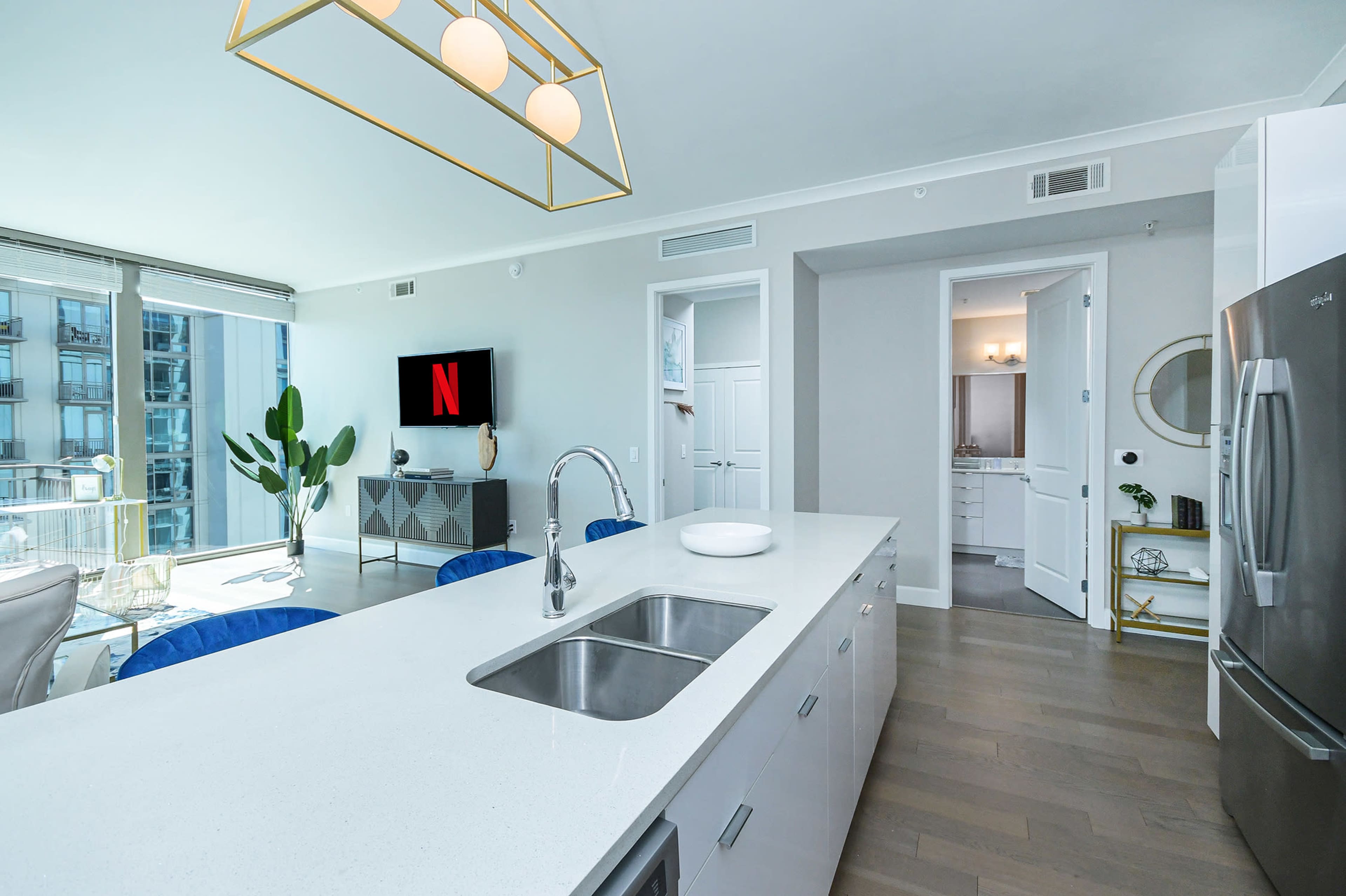 A modern kitchen with a white countertop, stainless steel appliances, and a large window displaying a city view, featuring a Netflix logo on a wall-mounted screen.