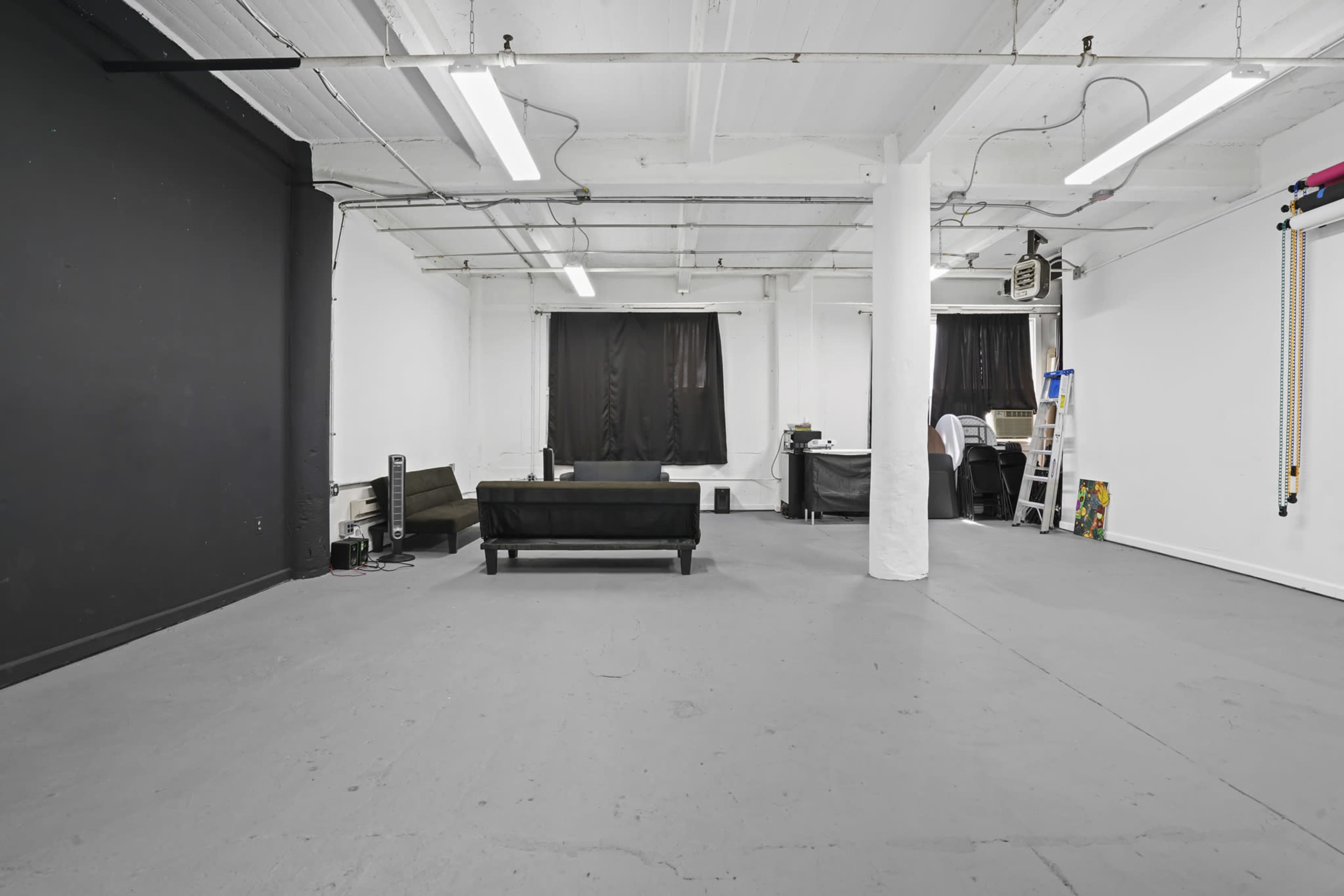 A spacious, industrial-style room with a gray floor, black and white walls, two black curtains, and minimal furnishings, including a black couch and a work area in the background.