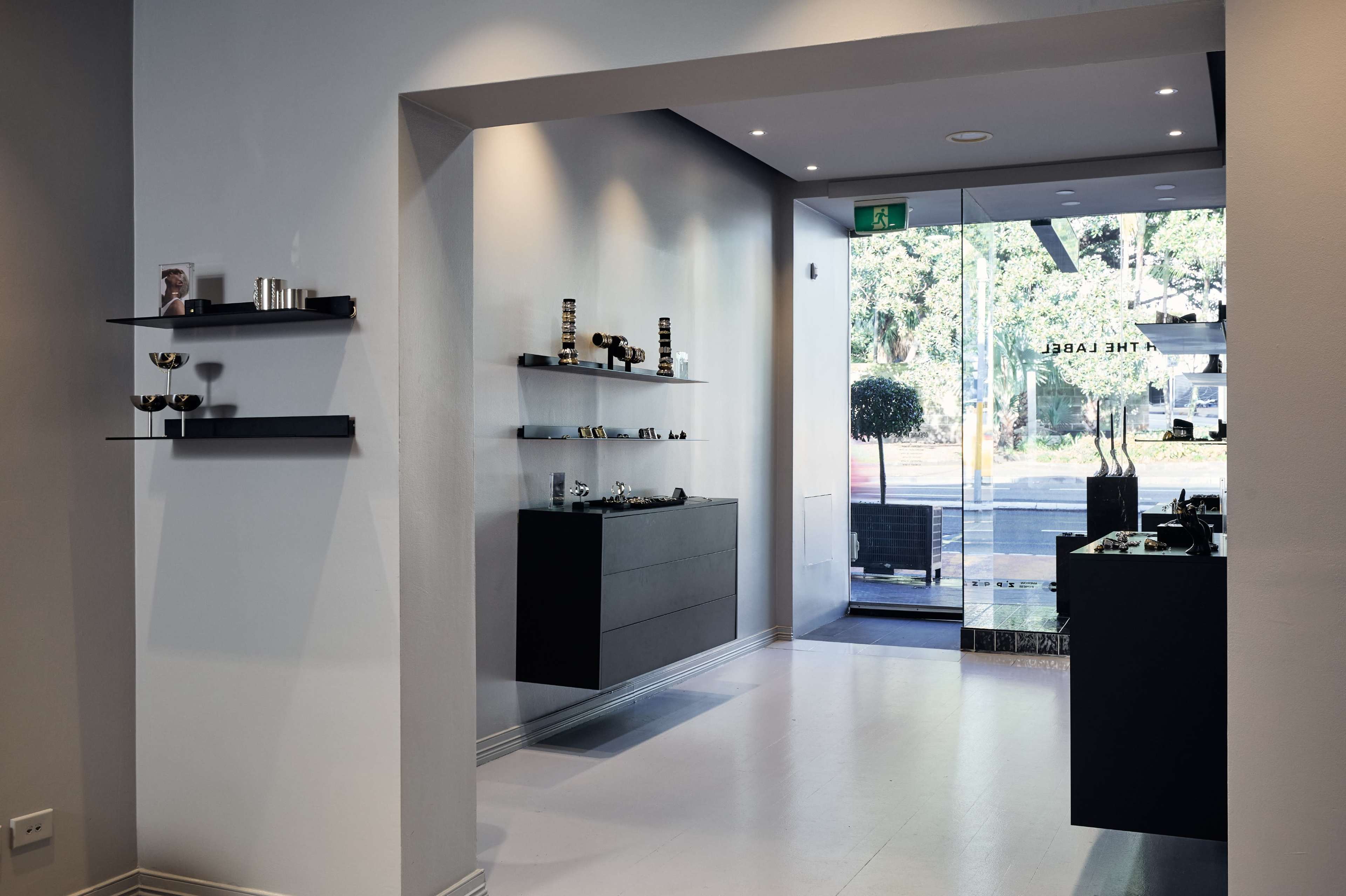 The image shows a modern boutique interior featuring minimalist shelving with various items displayed on the walls and a glass entrance that opens to the outside.