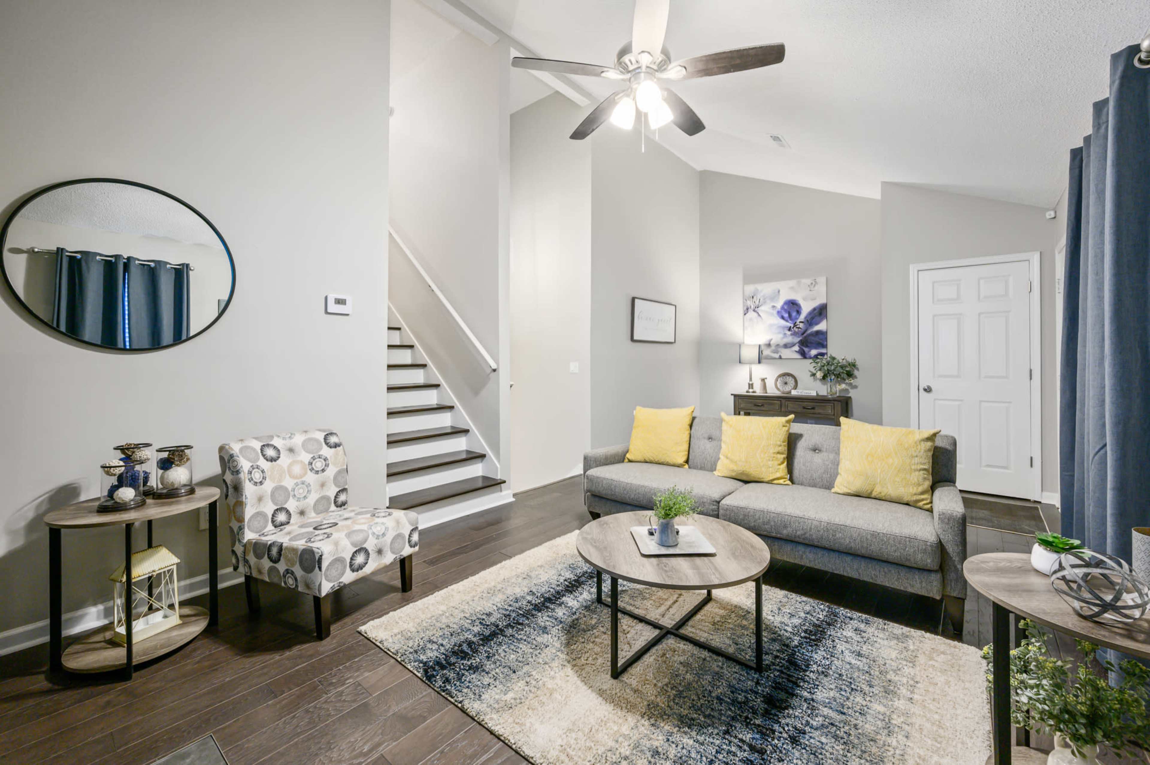 The image depicts a modern living room featuring a gray sofa with yellow cushions, a circular mirror, and a staircase leading to an upper level.