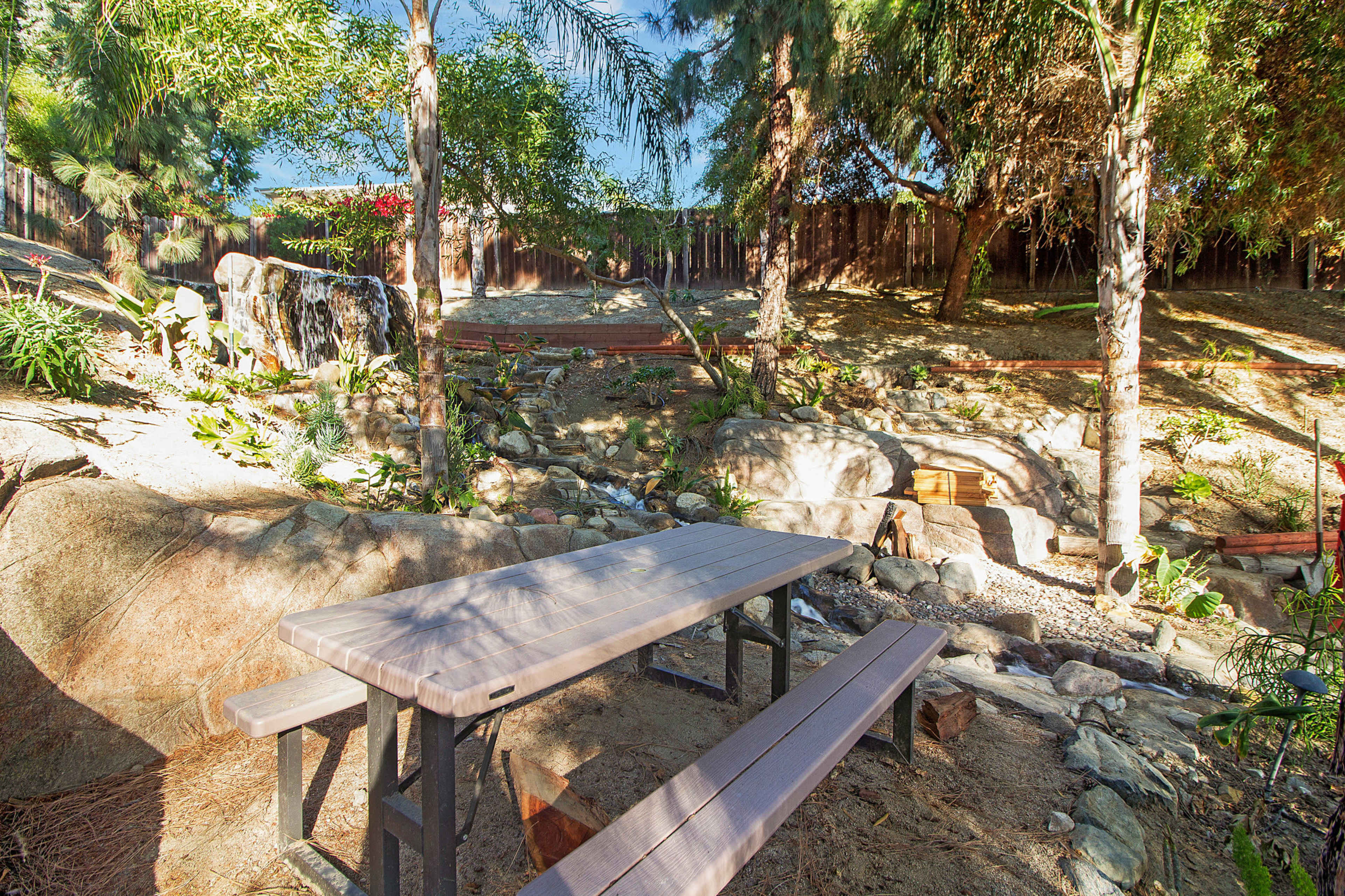A wooden picnic table is situated in a landscaped area surrounded by rocks, plants, and a small waterfall.