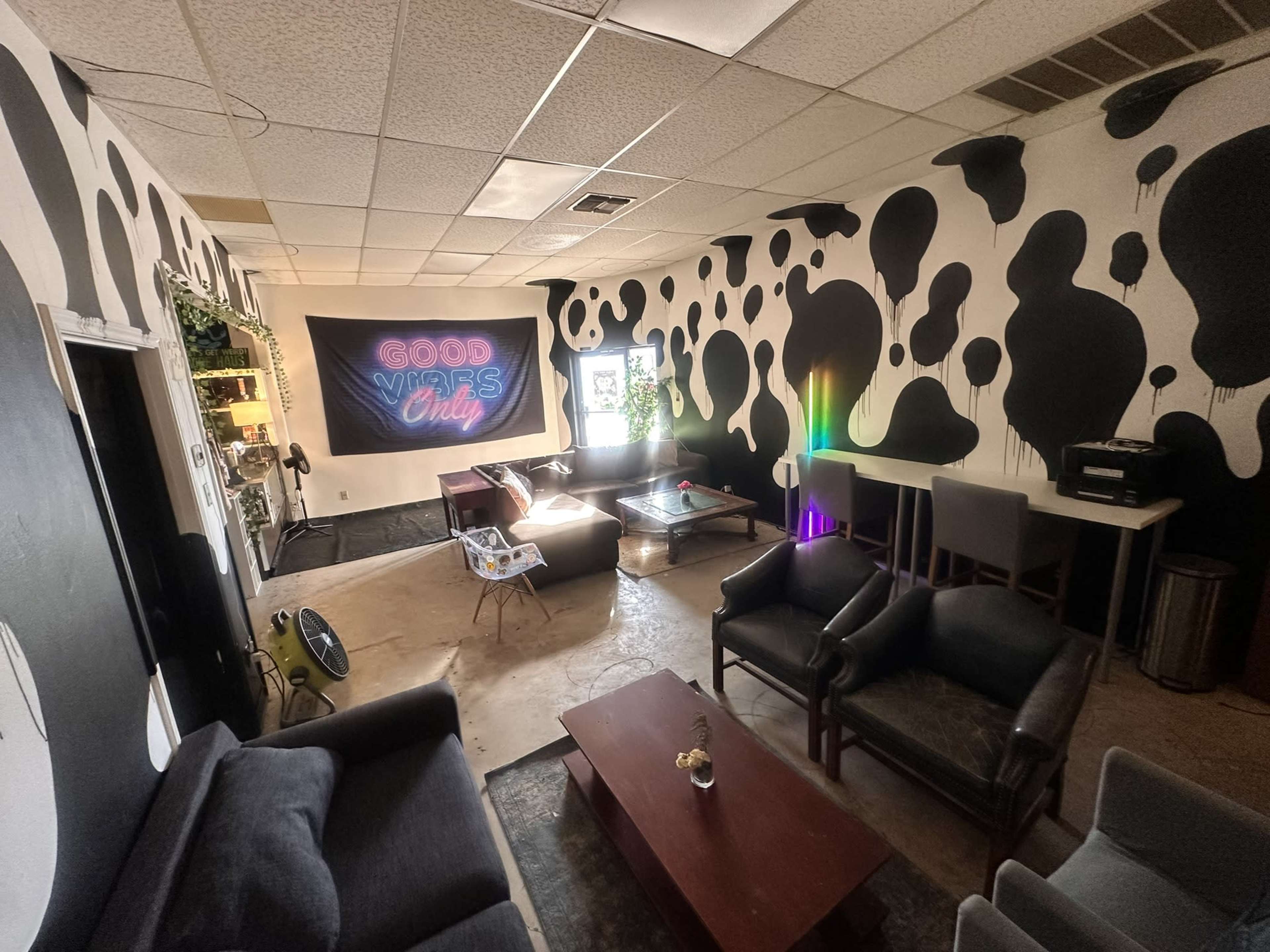The image shows an eclectic lounge with cow-patterned walls, comfortable seating, a table, and a neon sign that reads "Good Vibes Only."