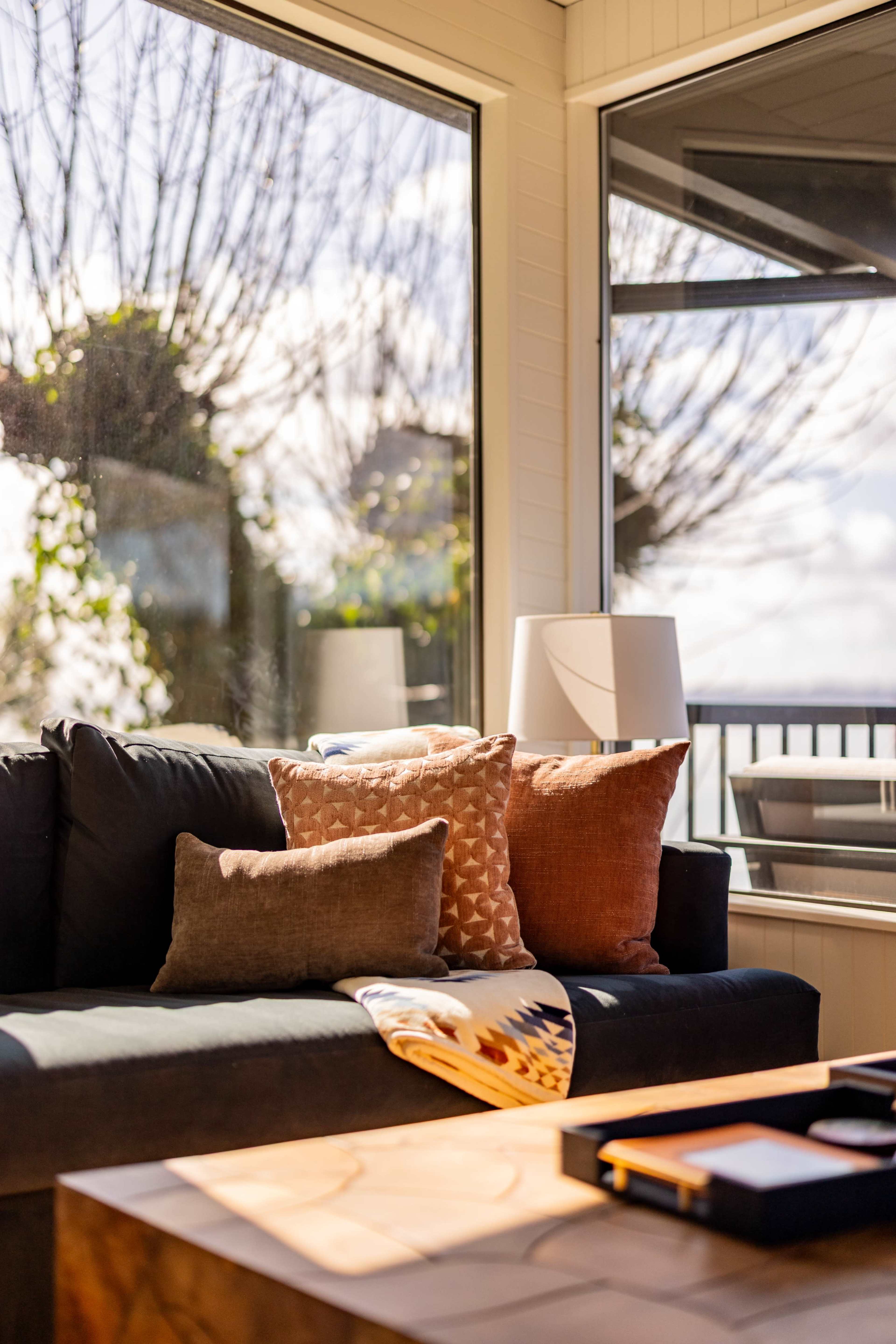 A living room features a sofa with decorative pillows, a wooden coffee table, and large windows showcasing a view of trees outside.