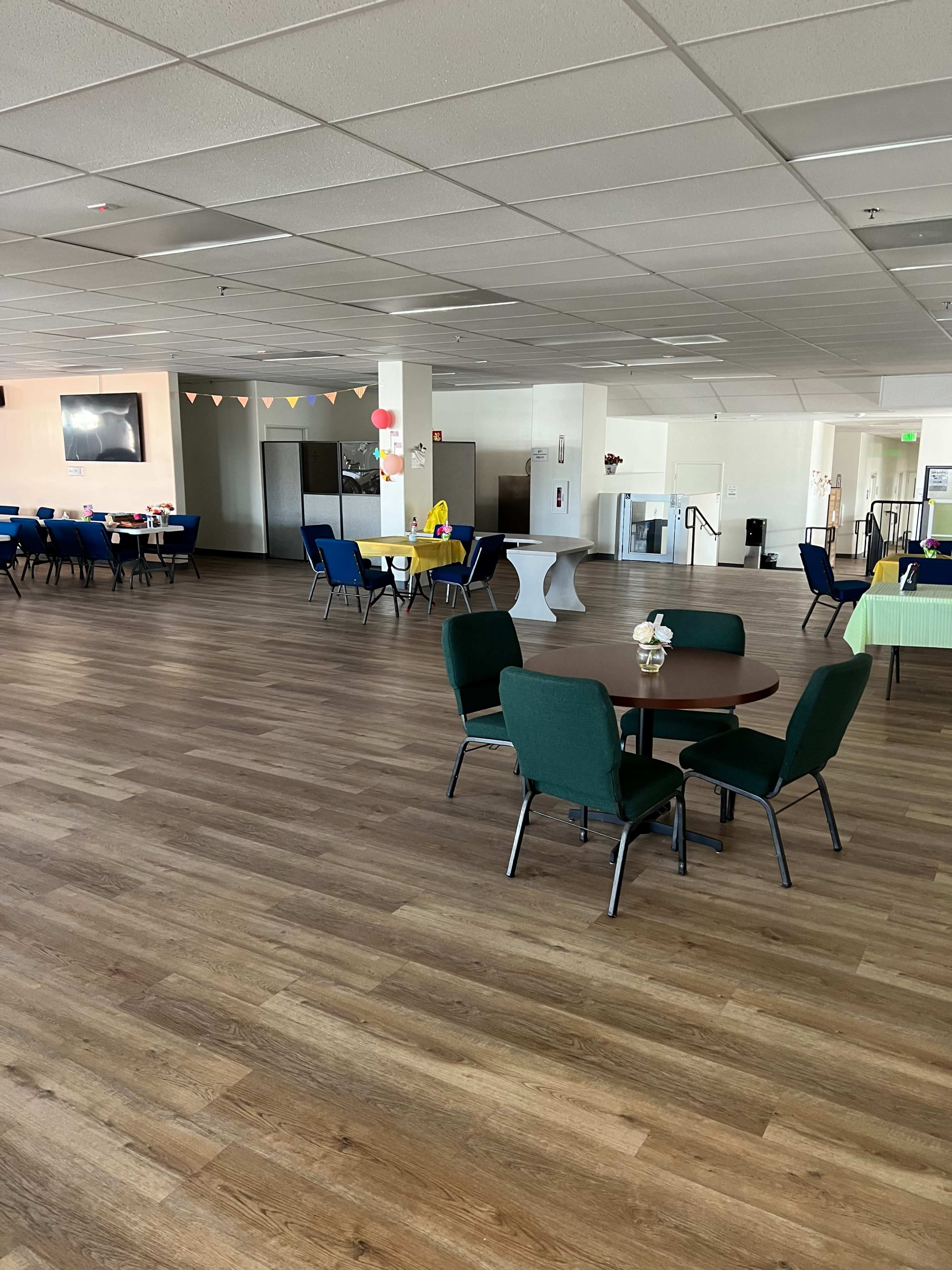 A spacious community hall with wooden flooring, several tables and chairs arranged in small groups, and a large empty area in the center.