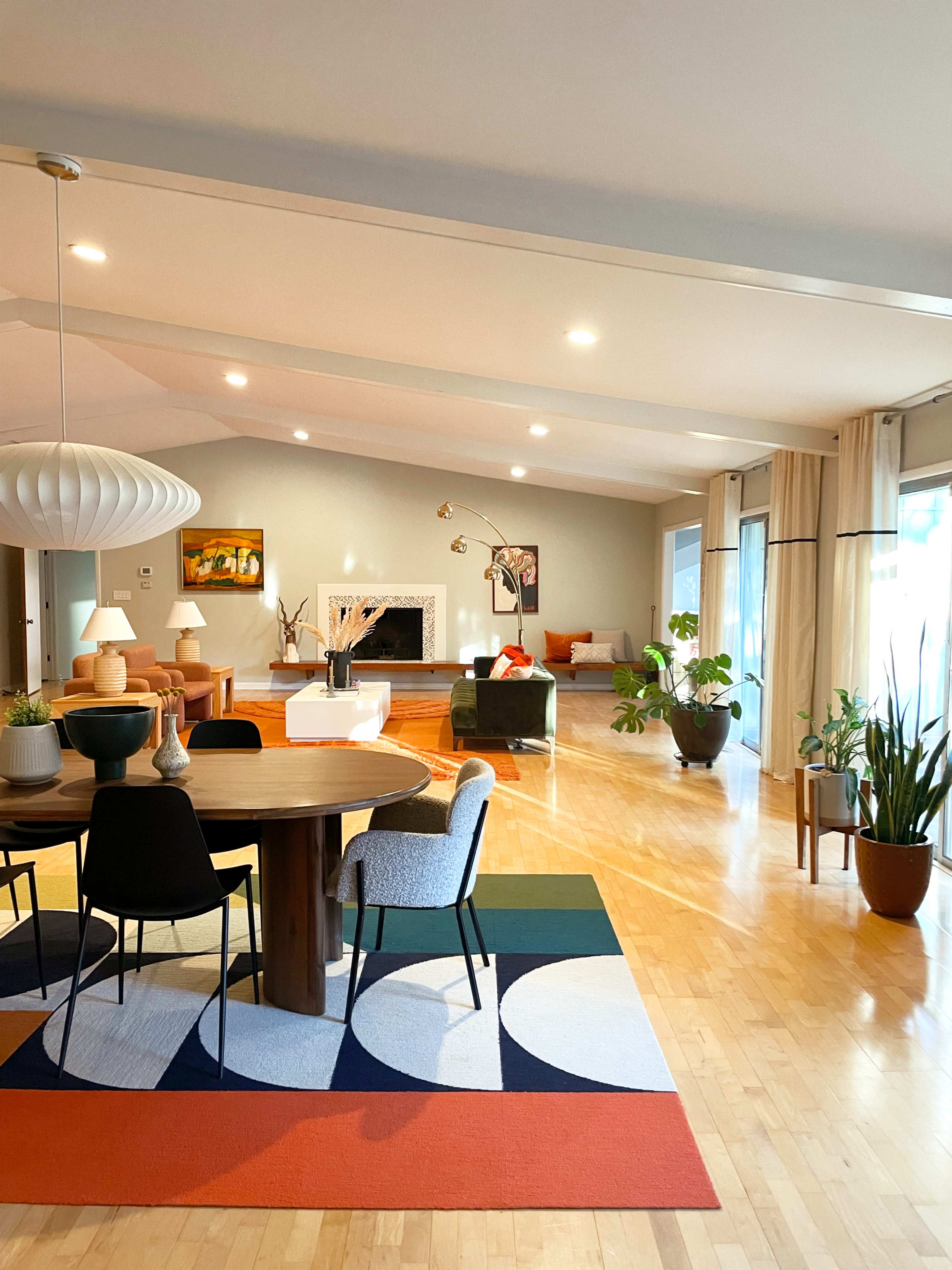 A spacious living area with a round dining table at the center, surrounded by modern chairs and a large area rug, featuring large windows and various indoor plants.